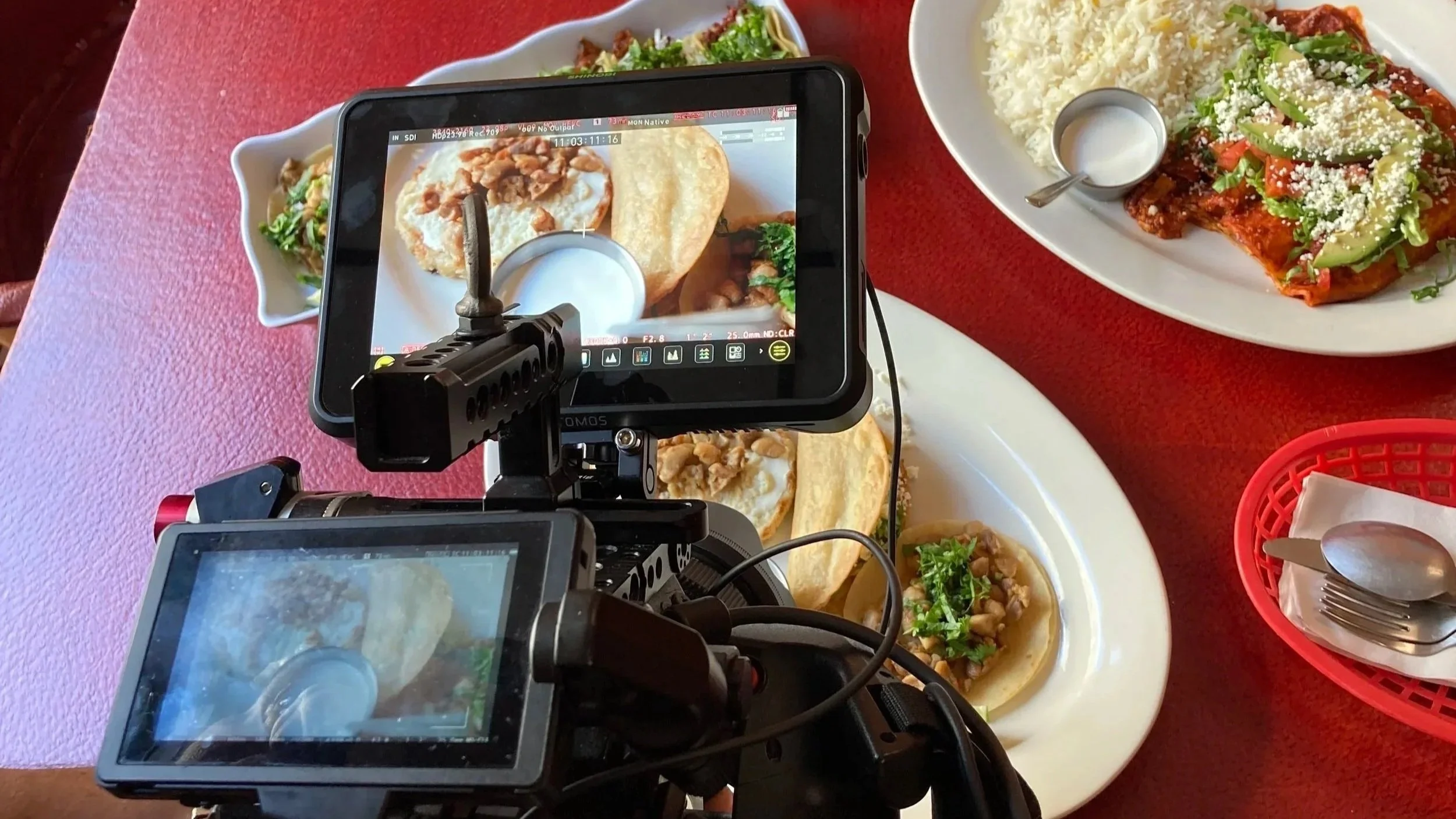 A camera filming food dishes on a red table, including plates with tacos, rice, and a dish topped with cheese and green peppers.