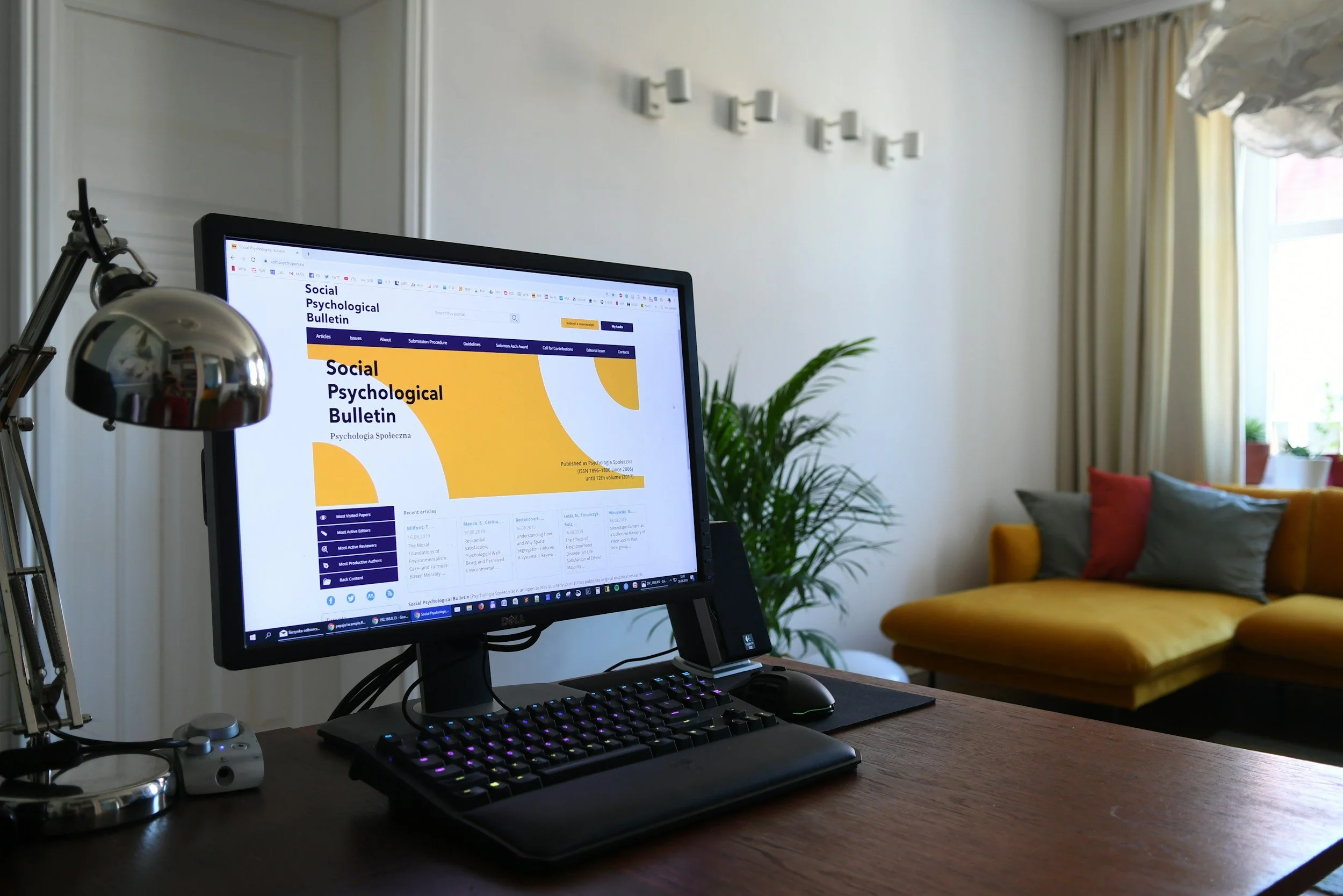 A computer monitor displaying a webpage, with a keyboard, mouse, and desk lamp in the foreground. A yellow chair with cushions and a window with curtains are in the background.