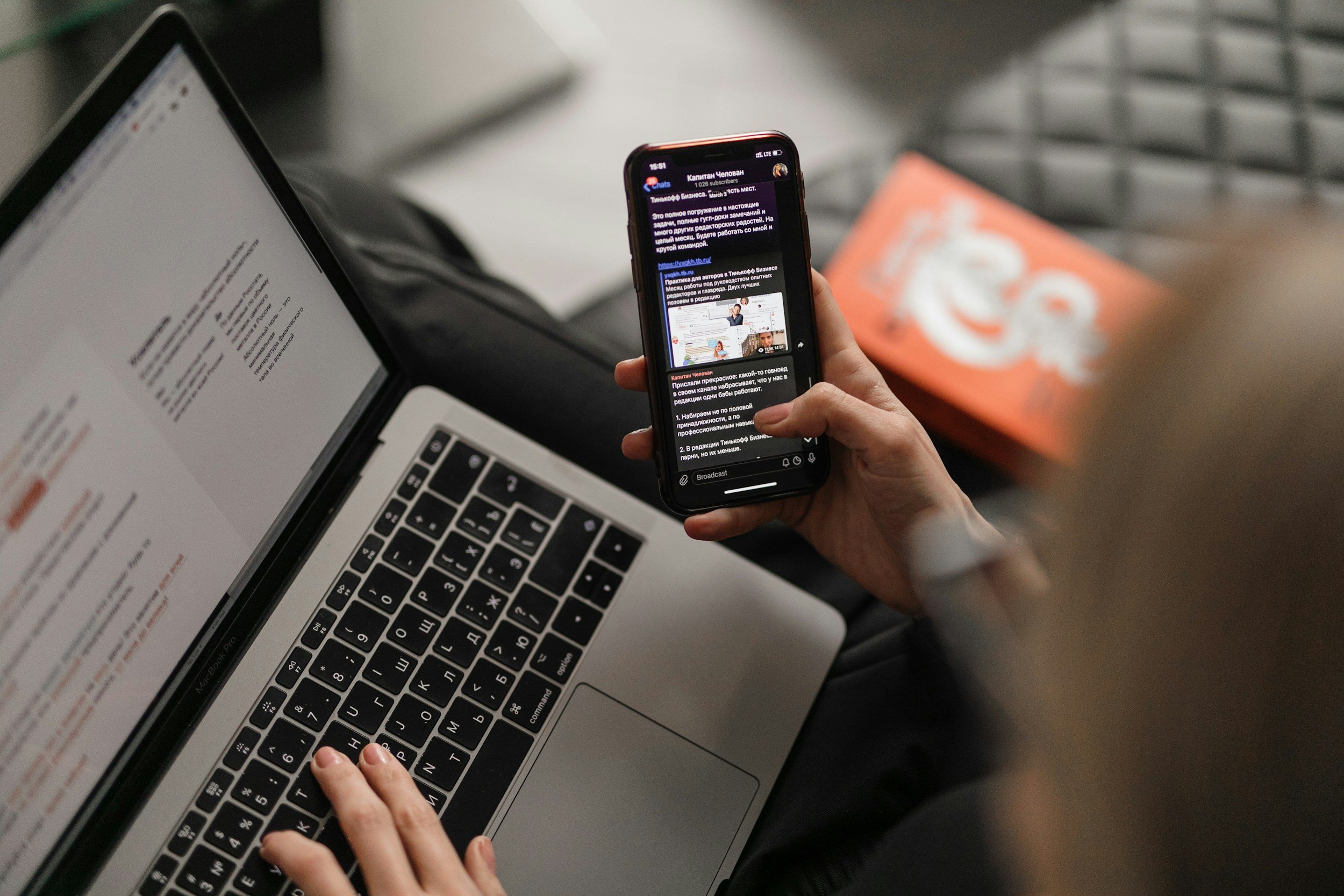 A person using a laptop computer and a smartphone. The laptop has a silver body and black keyboard, with some text on the screen. The person's hand is on the keyboard, and the smartphone displays a messaging app with text. There is a blurred orange box behind the phone and some out-of-focus background elements.