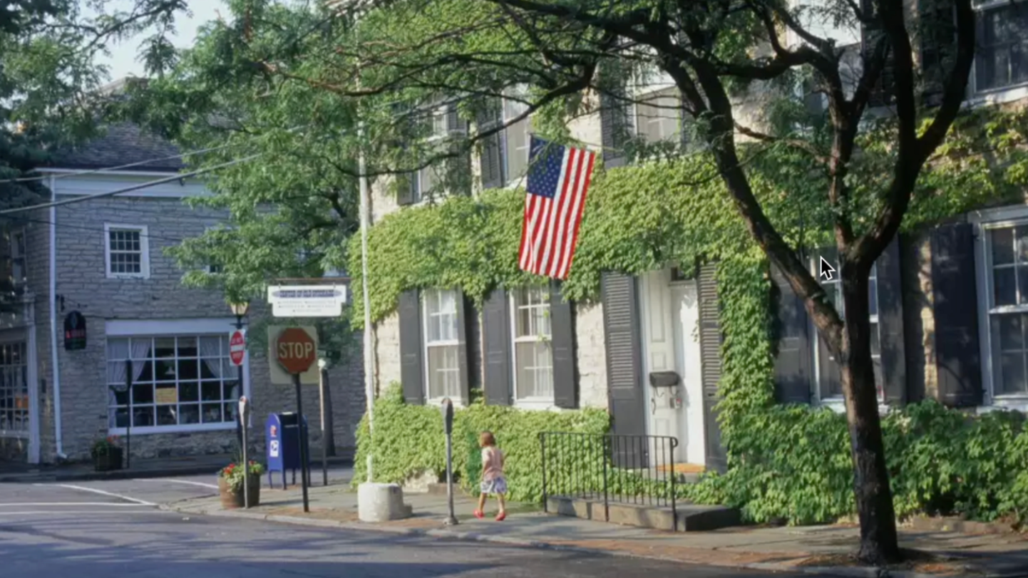 Street scene with a building covered in green ivy, American flag hanging, and a child walking on the sidewalk.