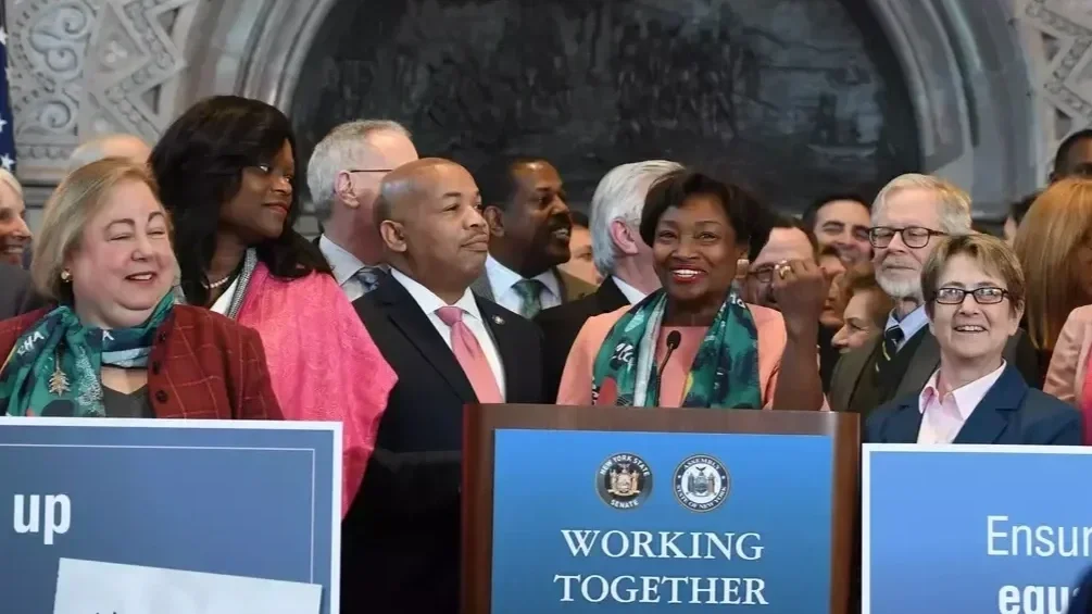 A diverse group of NY State legislators, including women and men of various ethnicities, standing behind a podium with a sign that says 'Working Together' at a formal event or press conference.