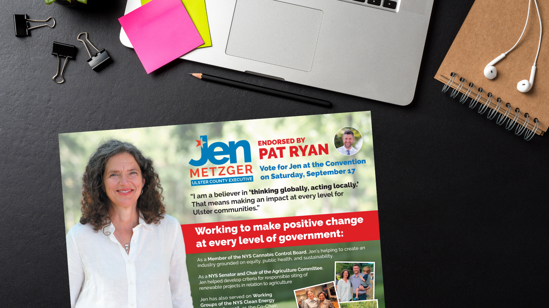A political campaign flyer endorsing Jen Metzger for Ulster County Executive, featuring her photo, a quote, and information about her work and endorsements, placed on a black desk with office supplies and a laptop.