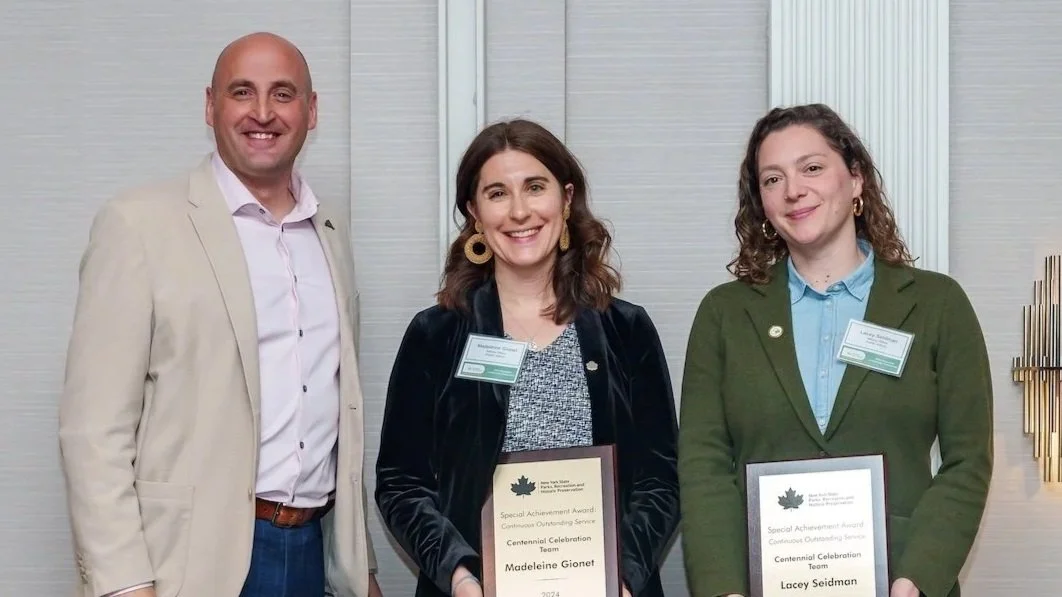 Three people standing indoors, with two women holding awards. The man on the left is wearing a beige blazer and pink shirt, the woman in the middle has dark hair and is wearing a black jacket with gold earrings, and the woman on the right has curly hair and is wearing a green blazer with a light blue shirt.