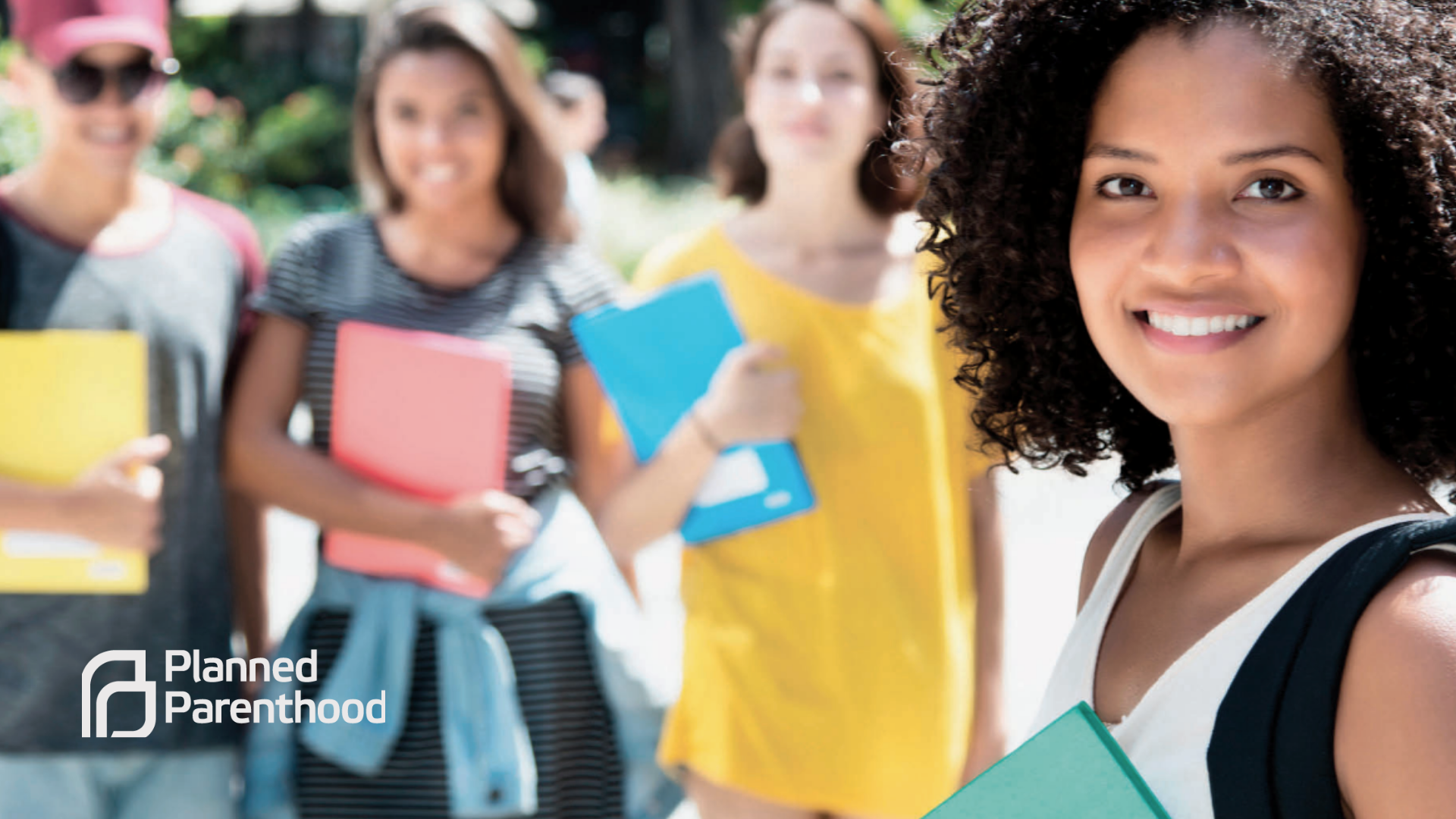 A group of diverse young women holding notebooks and smiling outdoors, with a logo for Planned Parenthood in the lower-left corner.