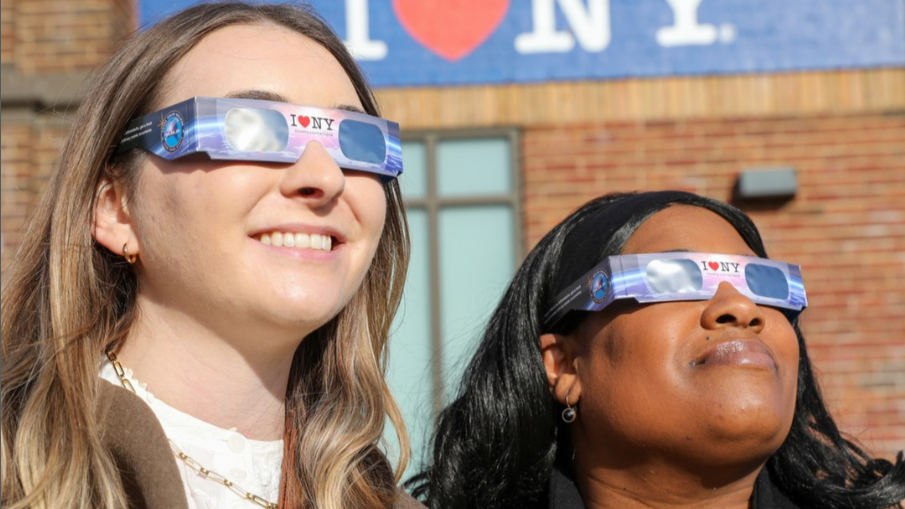 Two women wearing eclipse glasses in front of a brick building with a blue banner that says "I ♥ NY" in the background.