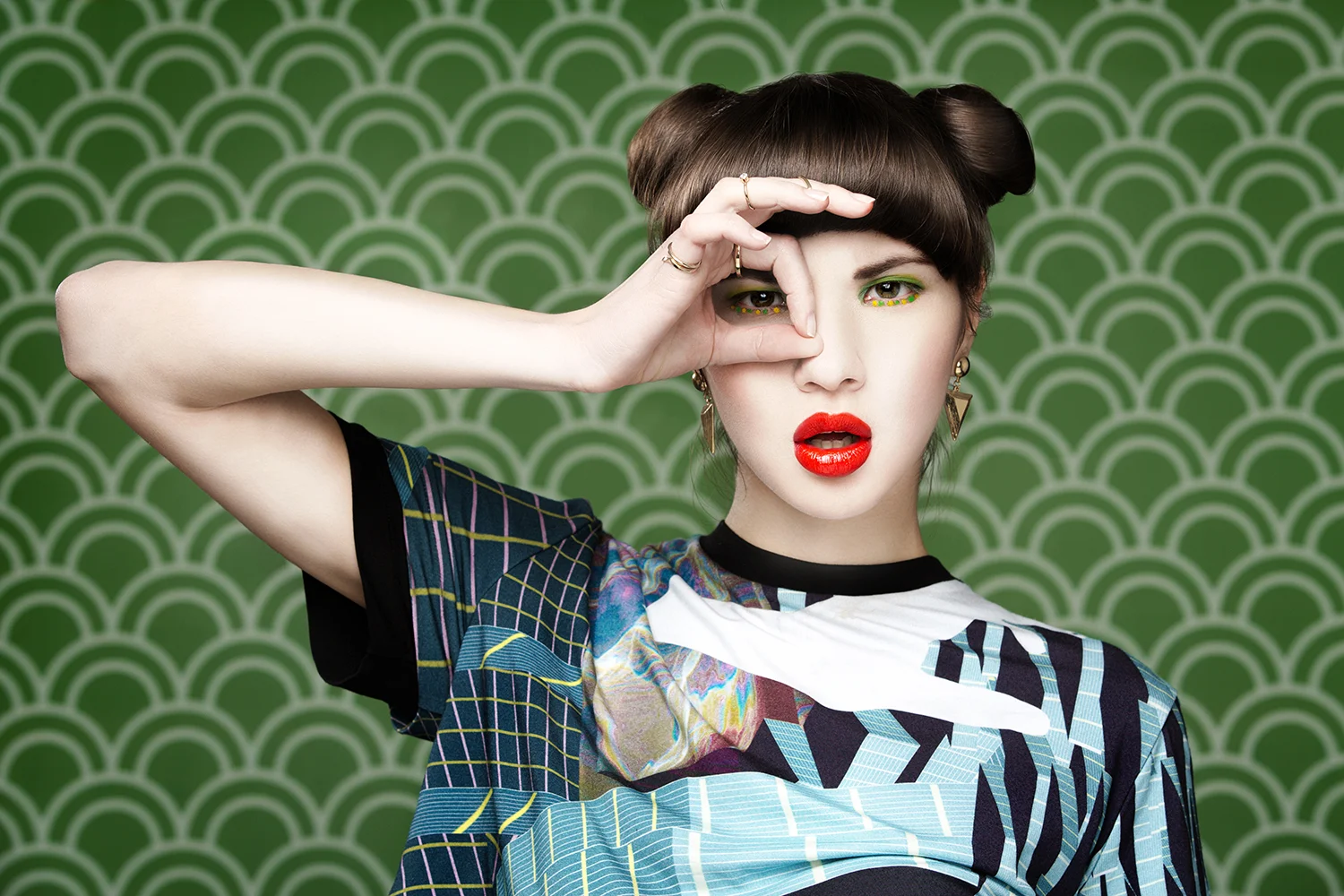 Ragazza con trucco vivace, capelli corti e vestito colorato, fa un gesto con la mano attorno all'occhio e guarda attraverso le dita, sfondo verde con motivo a onde