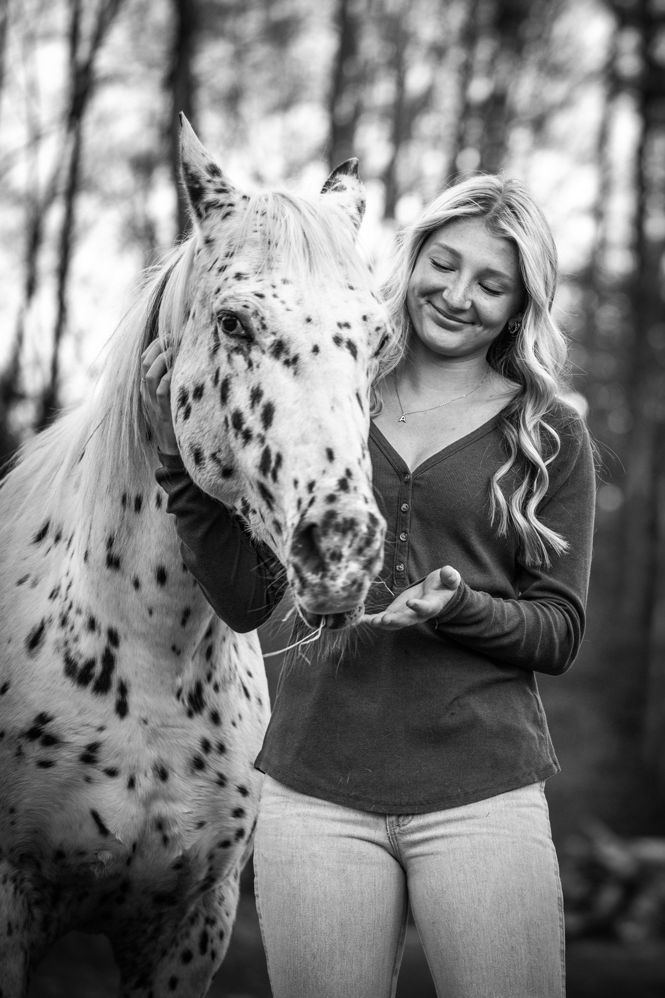 A black and white photo of a smiling woman with long hair standing next to a dappled horse in a forest setting. She is gently holding the horse's mane and offering food in her hand.