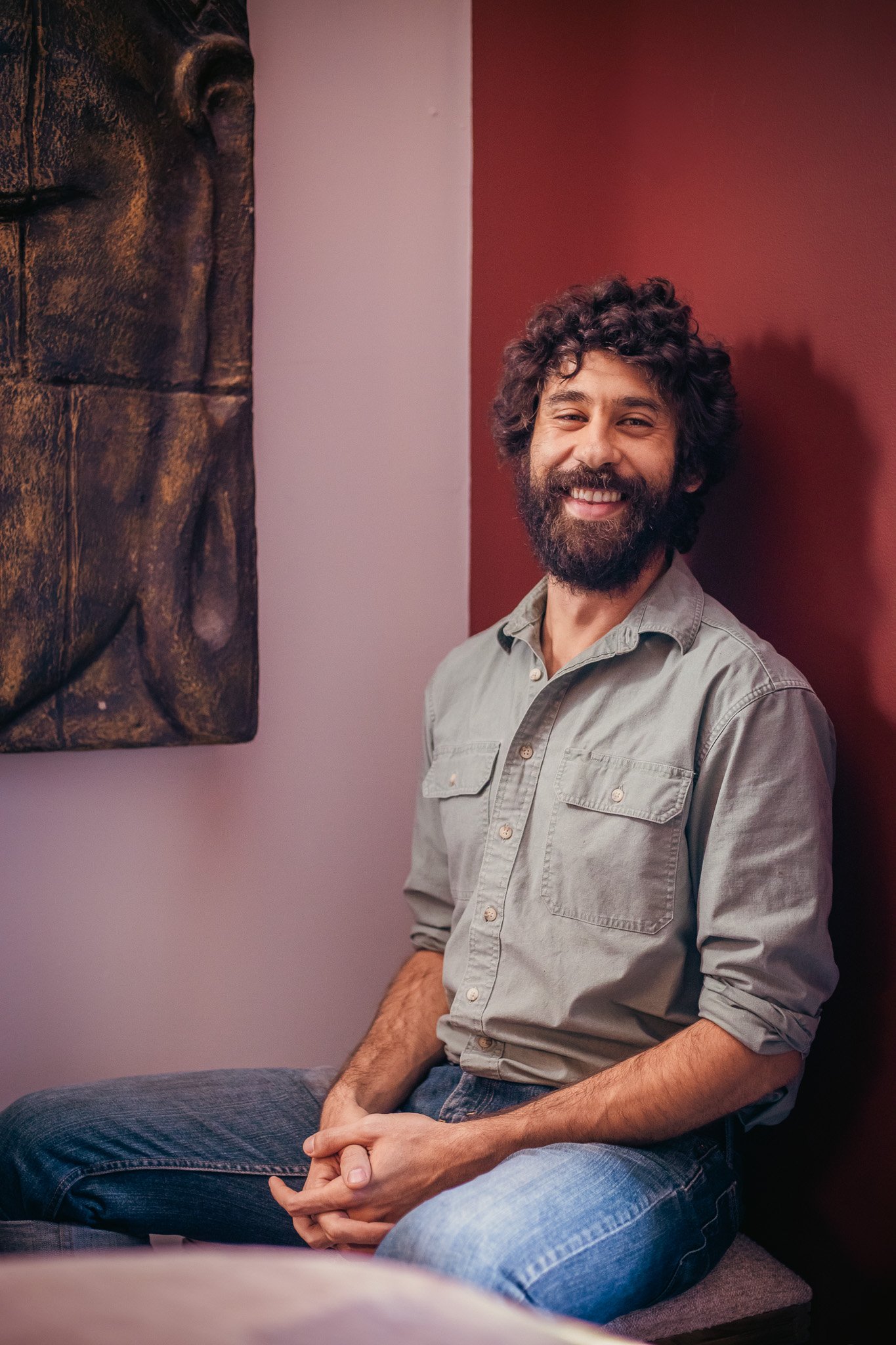 Bearded man smiling, sitting against a red wall, wearing a green shirt and jeans.