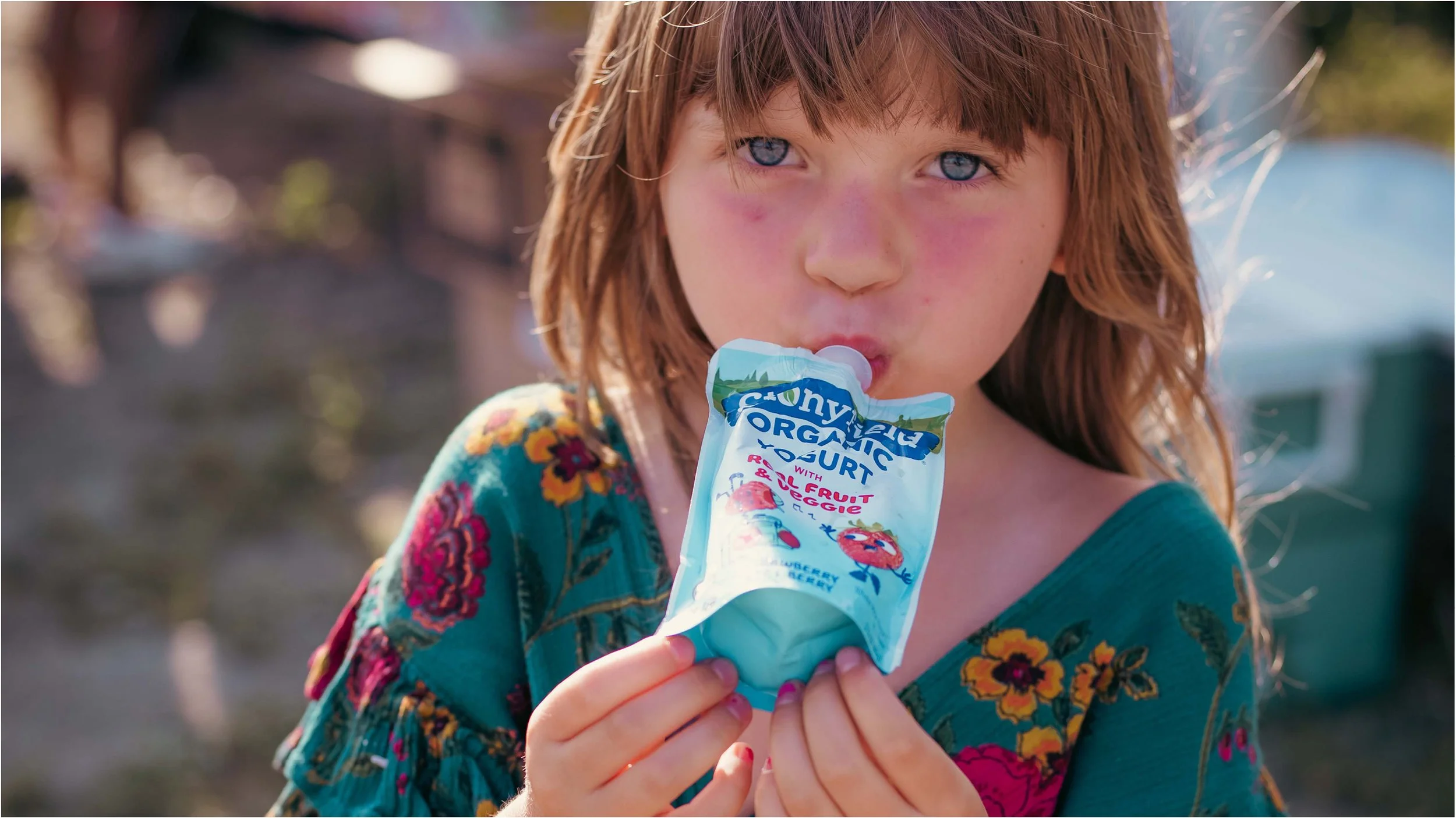 A young girl with red hair and blue eyes holding and about to eat a yogurt pouch outdoors.