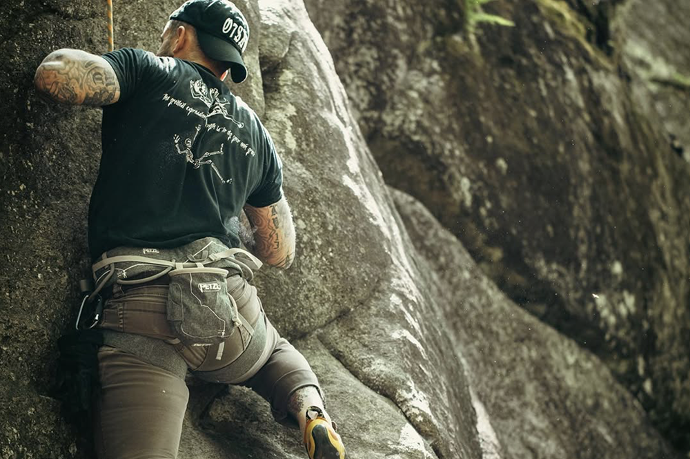A man with tattoos wearing a black cap, black shirt, and beige pants is rock climbing on a rugged outdoor cliff.