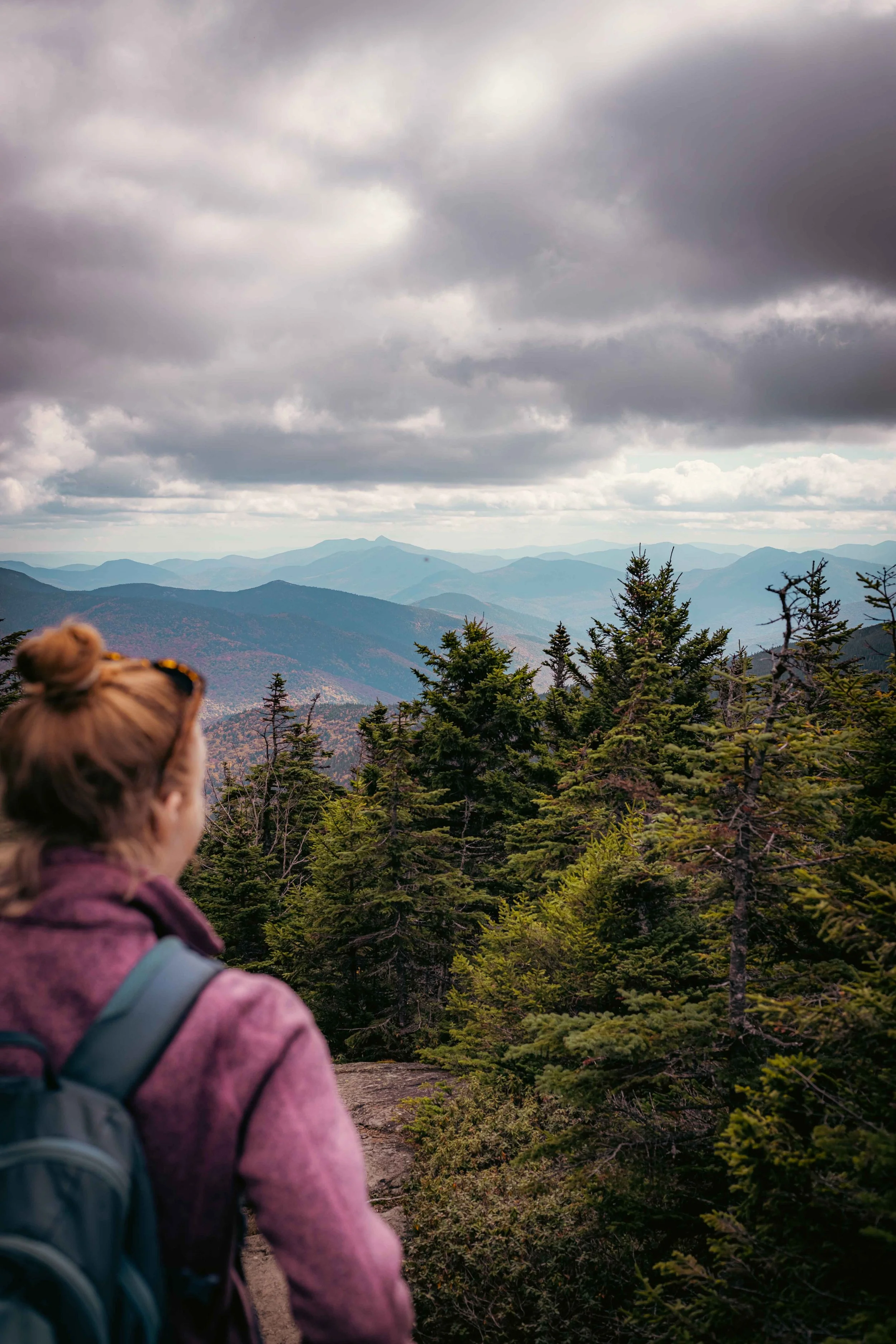 A woman with a backpack hiking in a mountain forest, looking at distant mountain ranges under cloudy skies.