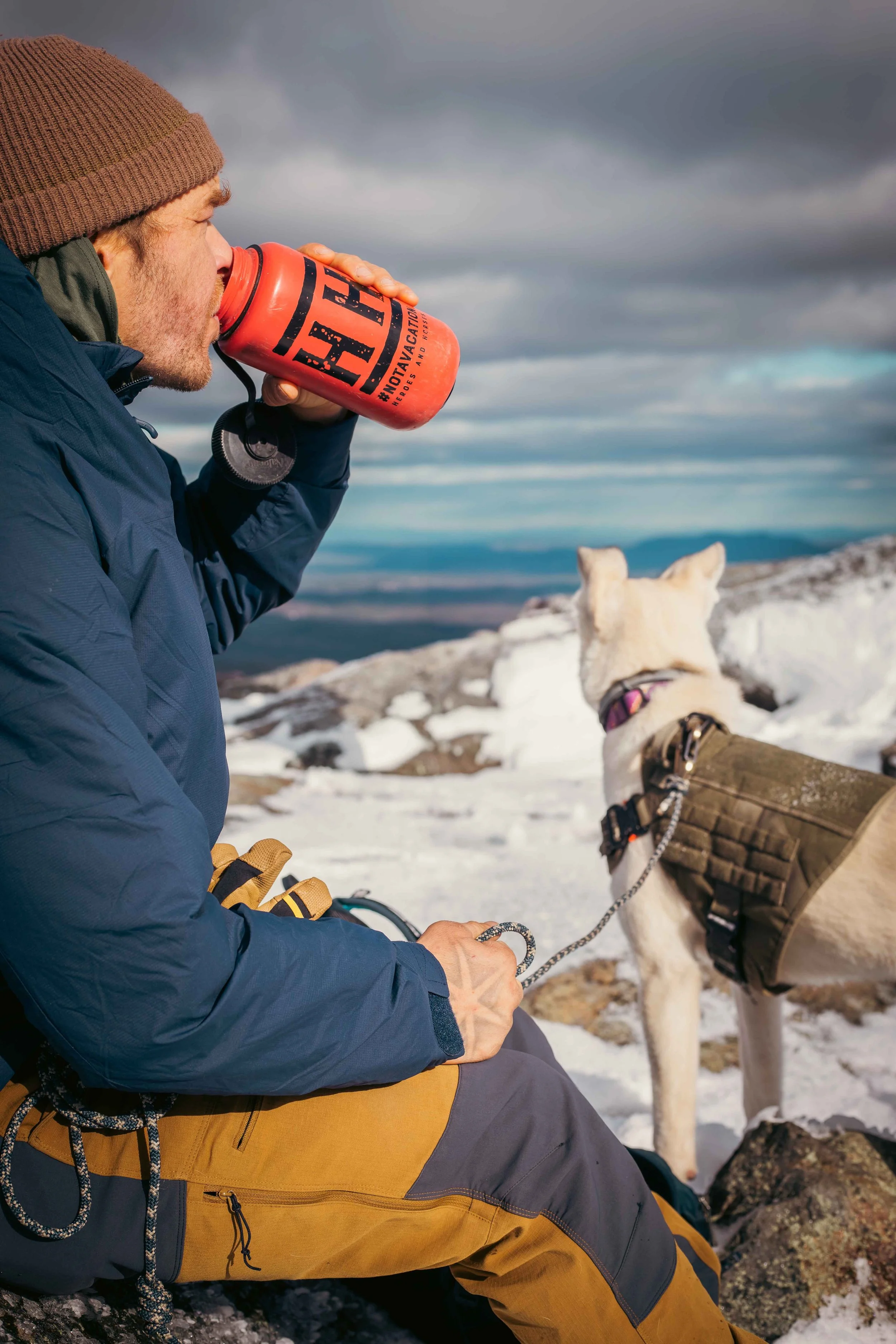 A person in winter clothing drinks from an orange water bottle while holding a dog on a leash in a snowy mountain landscape.