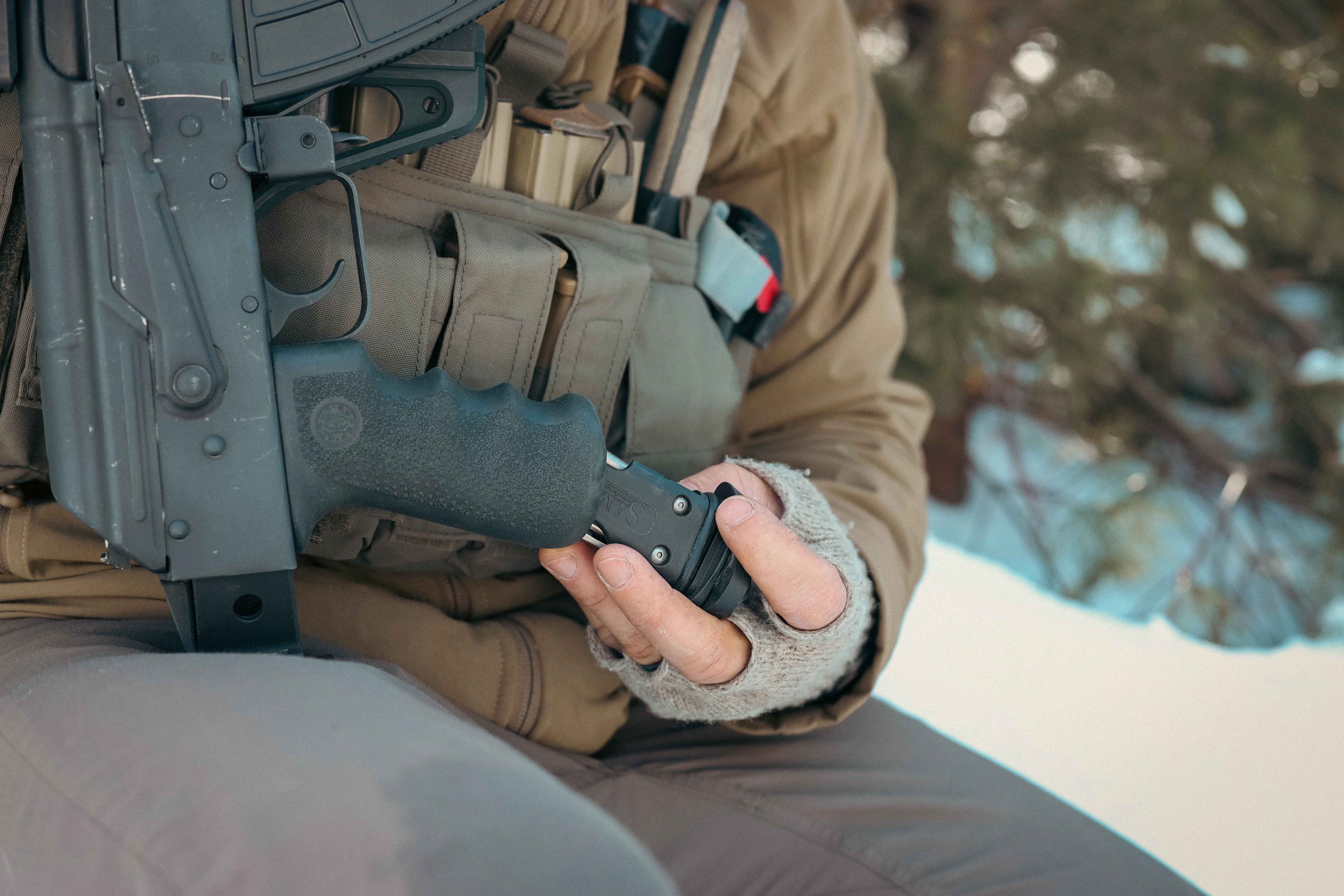 Person holding rifle and knife, wearing tactical gear in snowy outdoor setting.