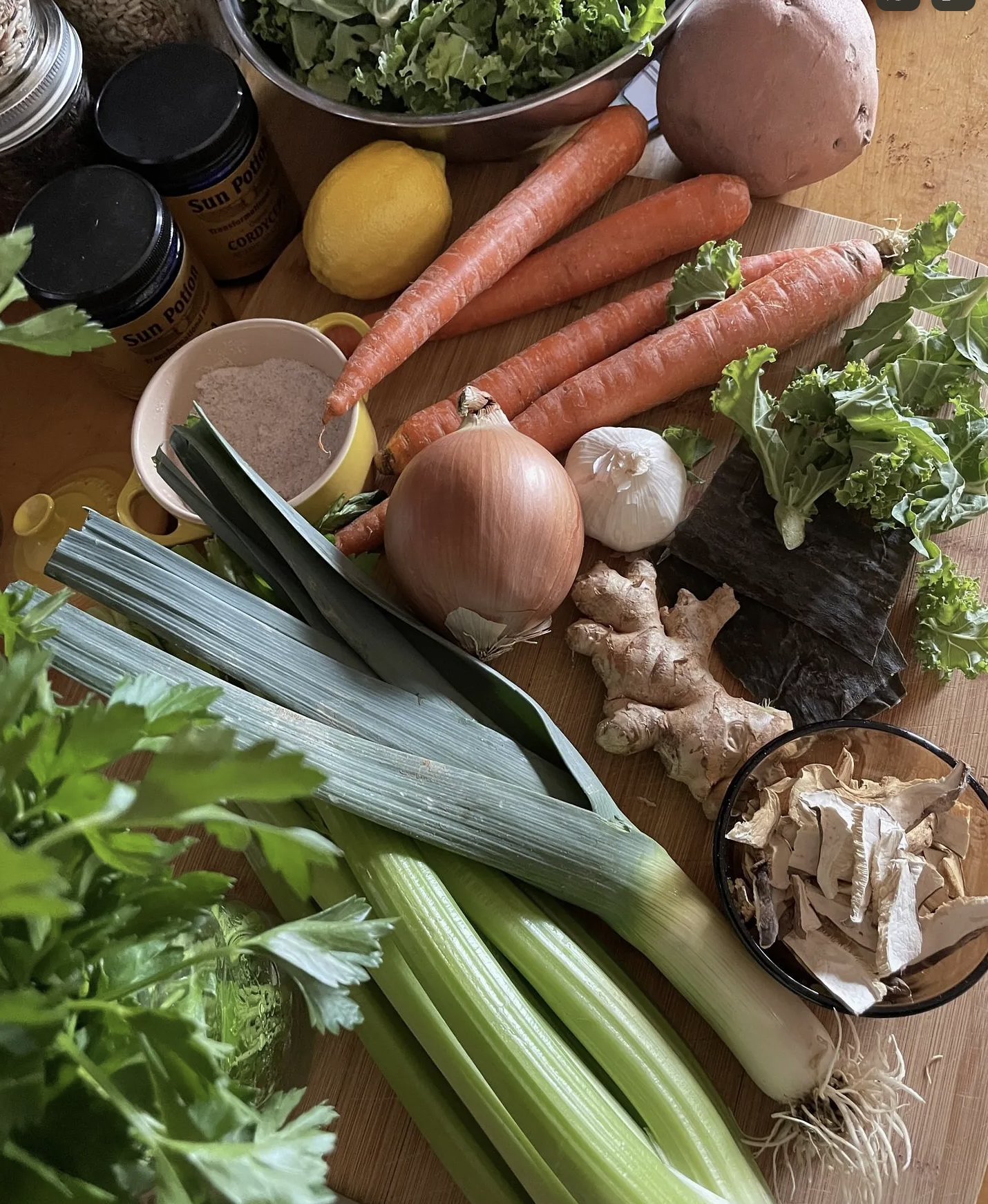 A variety of fresh vegetables including carrots, sweet potatoes, garlic, onion, ginger, leek, and greens on a wooden surface, with jars of supplements and a bowl of chopped greens in the background.