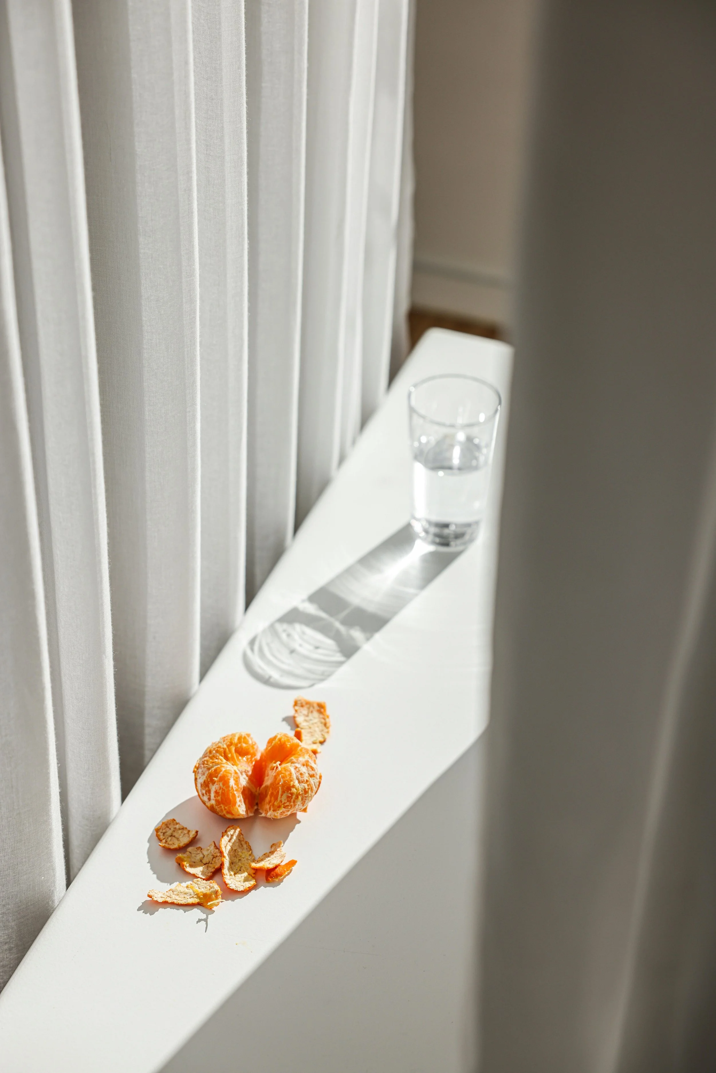 A white windowsill with a glass of water, a peeled orange, and orange segments, with sunlight casting shadows.