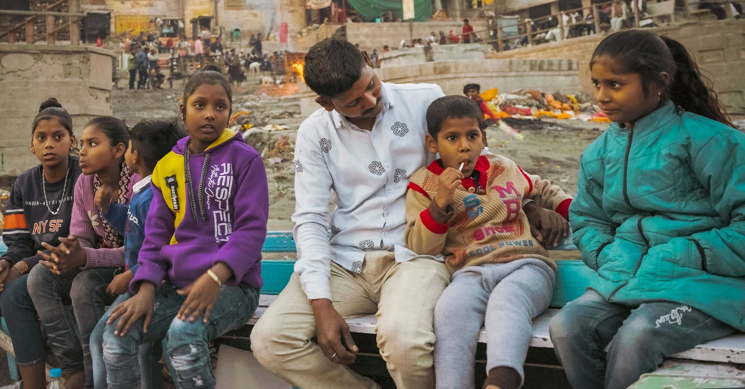 Group of children and a man sitting on a bench outdoors in a crowded area, some children wearing colorful jackets and sitting with various expressions.