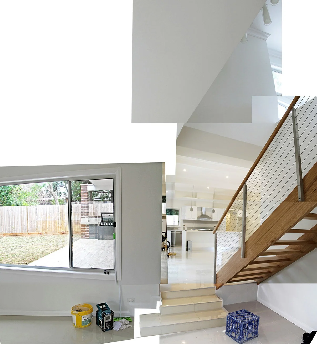 Interior of a modern home with white walls and flooring, a staircase with wooden steps and metal railing, a large window revealing a backyard, and kitchen area in the background.