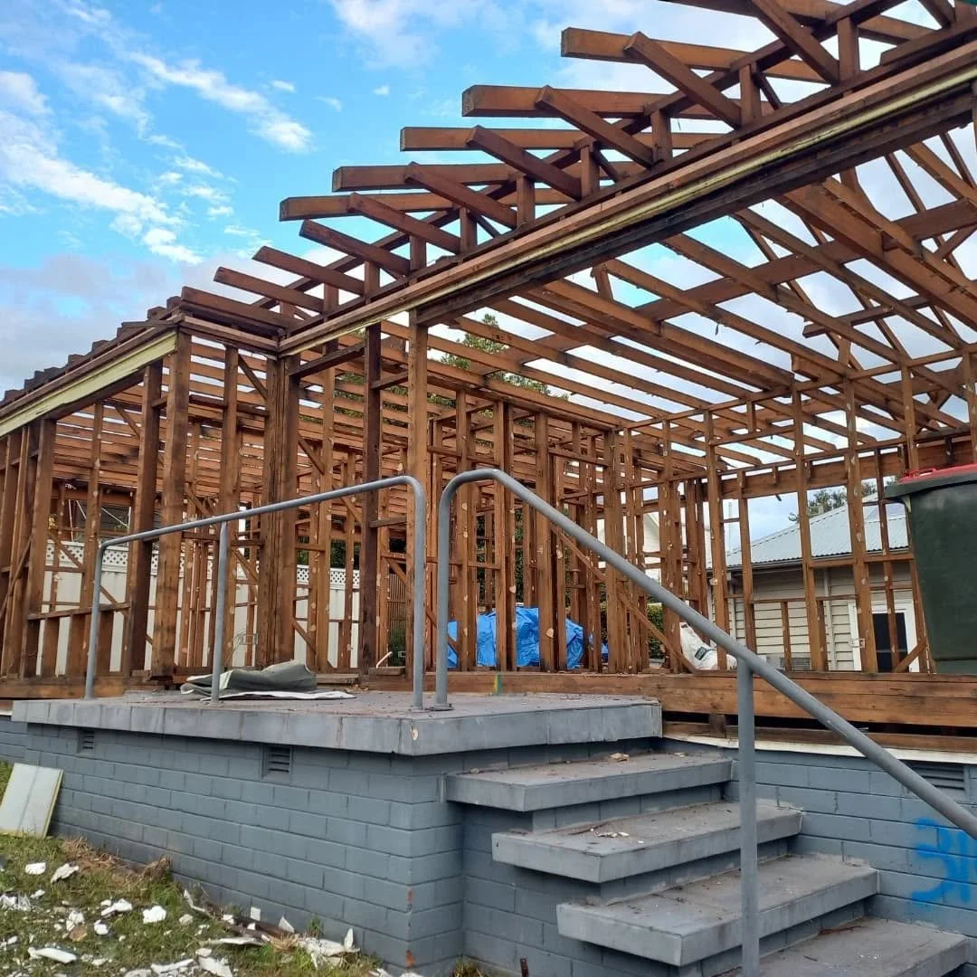 Wooden house frame under construction on a concrete block foundation with stairs, set against a partly cloudy sky.