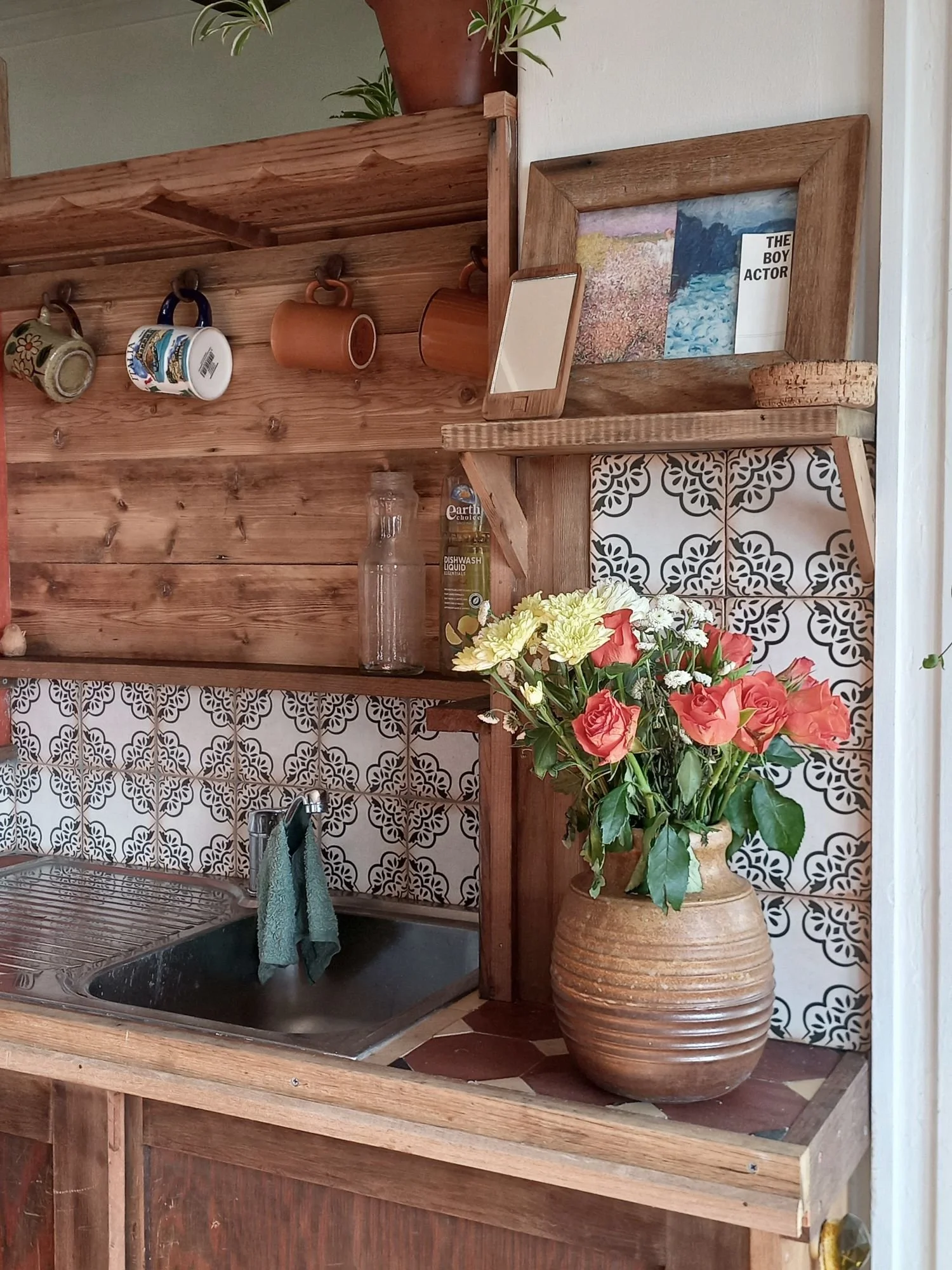 A kitchen area with a wooden countertop, hanging coffee mugs, a glass jar, and a large vase with a bouquet of pink and white flowers. There is a decorative tiled backsplash and a framed picture with landscape art.