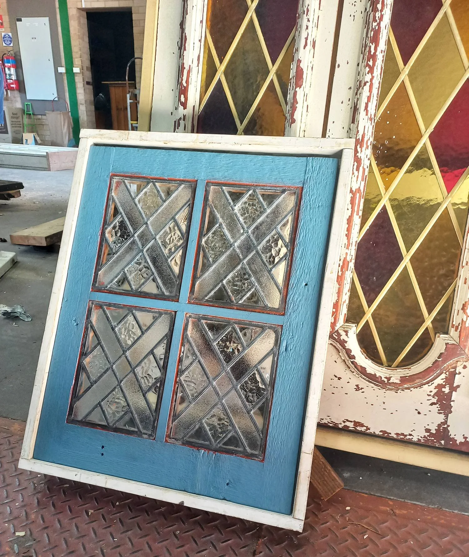 Two vintage stained glass and painted wood doors, one blue with geometric glass panels and the other with a distressed paint finish and diamond-shaped colored glass panels, leaning against each other in a cluttered workshop.