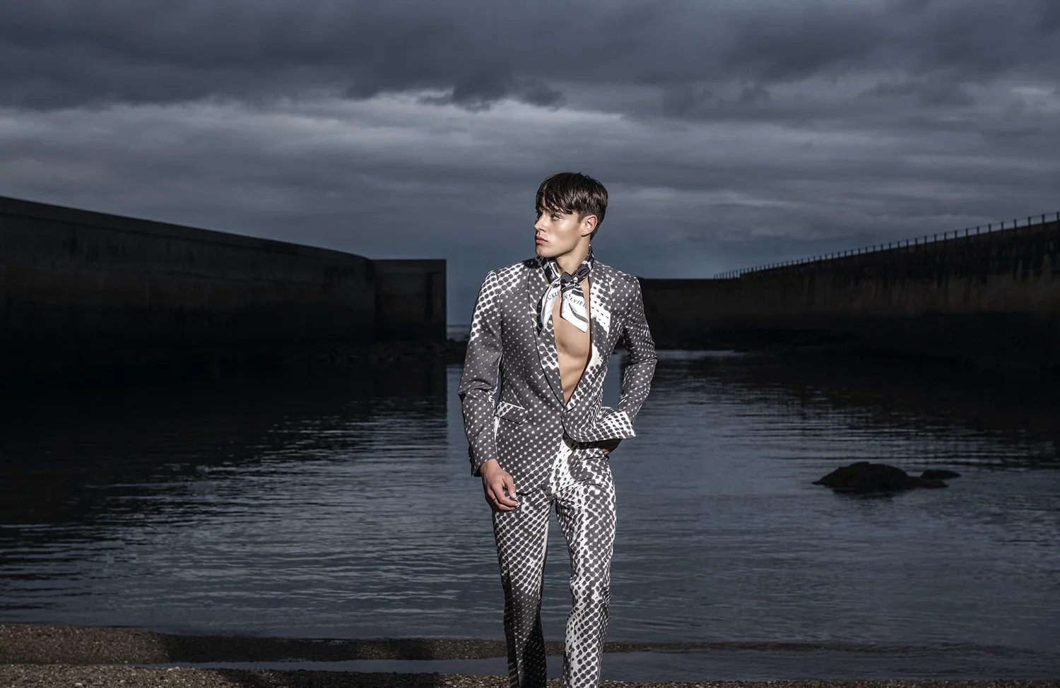 A model in a silver-patterned Miguel Vieira suit, photographed beside a concrete breakwater under heavy cloud