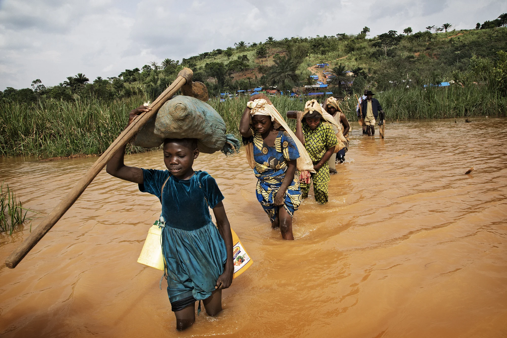 Miners and Traders cross the flooded river to the Gold mine in Baru, Mongbwalu. Recently discovered it has over 3000 miners working there extracting gold. Many of them are soldiers who have deserted and come to look for money. Whole families have relocated to be close to the mine. Children are used extensively to transport the gold from the mine, often carrying 25 -40 kilos.
