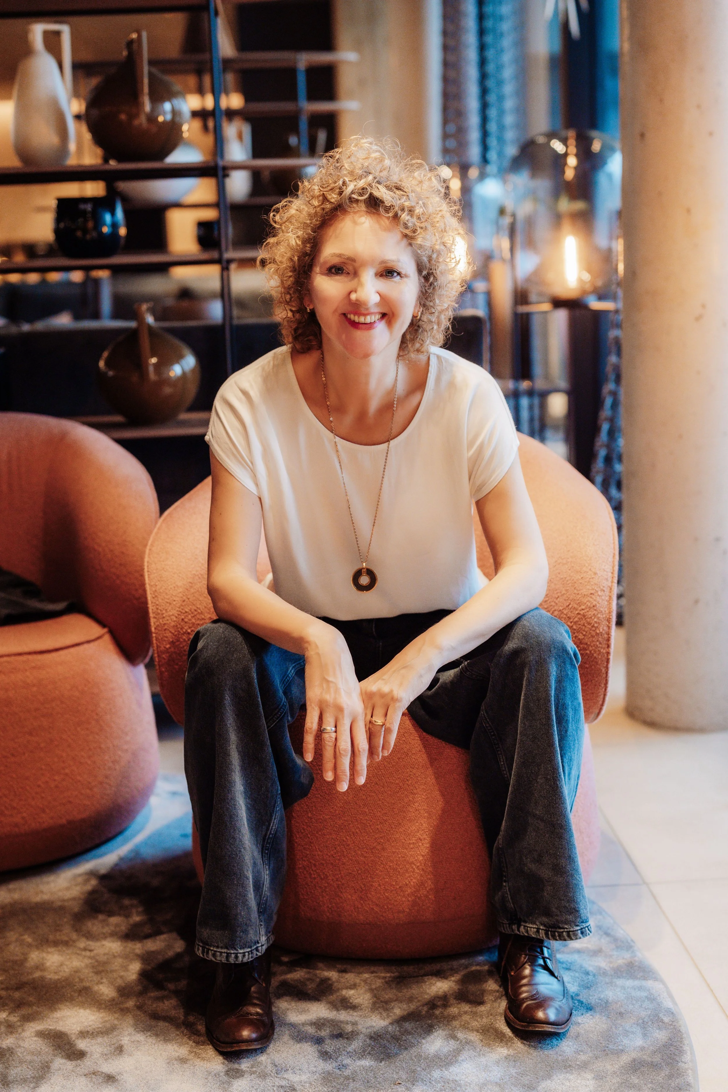 Monika sitzt ruhig auf einem Sessel, leicht nach vorne geneigt. Im Hintergrund ein Regal mit braunen Vasen und warmes, indirektes Licht.