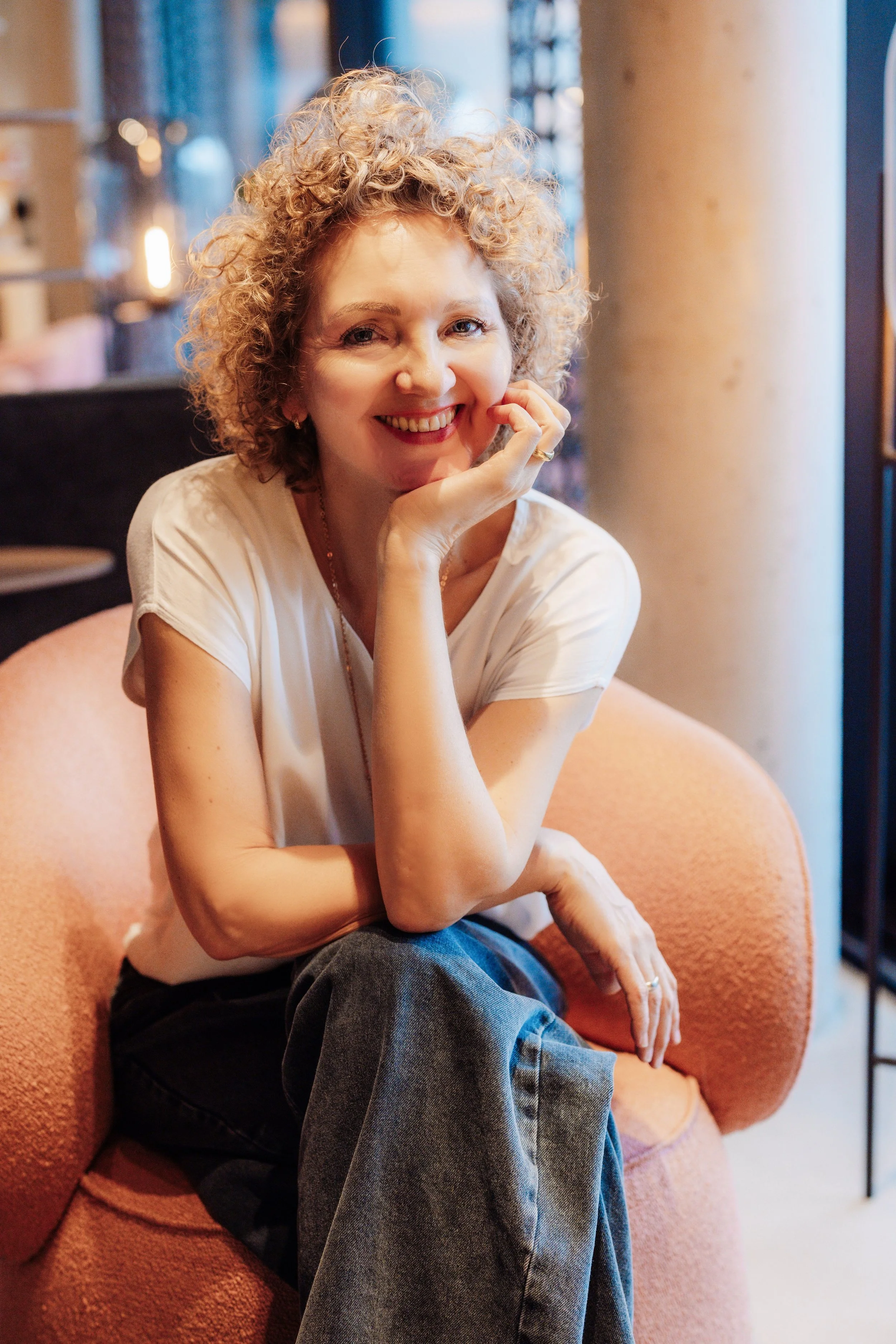 Monika sitzt ruhig auf einem Sessel und lächelt. Im Hintergrund ein Regal mit braunen Vasen und warmes, indirektes Licht.