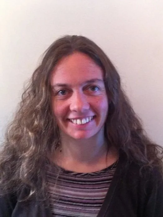 A woman with long, curly, brown hair smiling at the camera, wearing a striped shirt and black jacket, against a plain light-colored background.