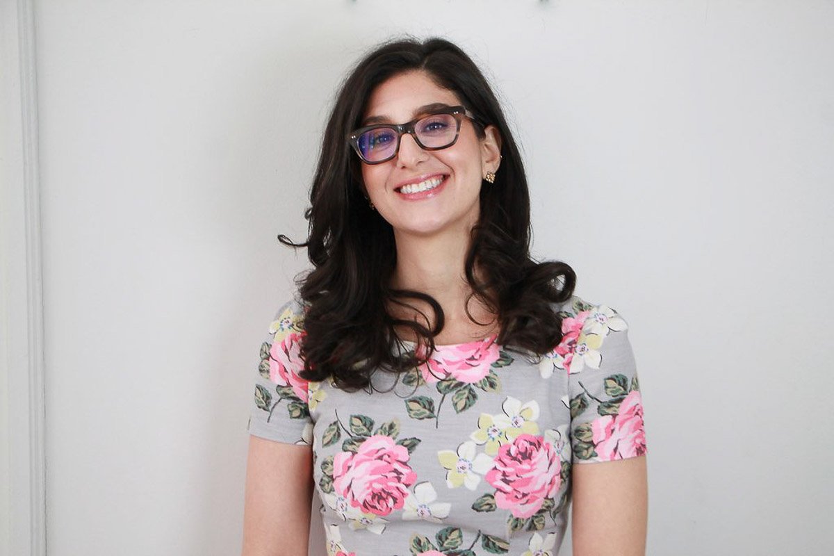 Holistic therapist smiling warmly, wearing a floral top and glasses, standing against a soft, light background.