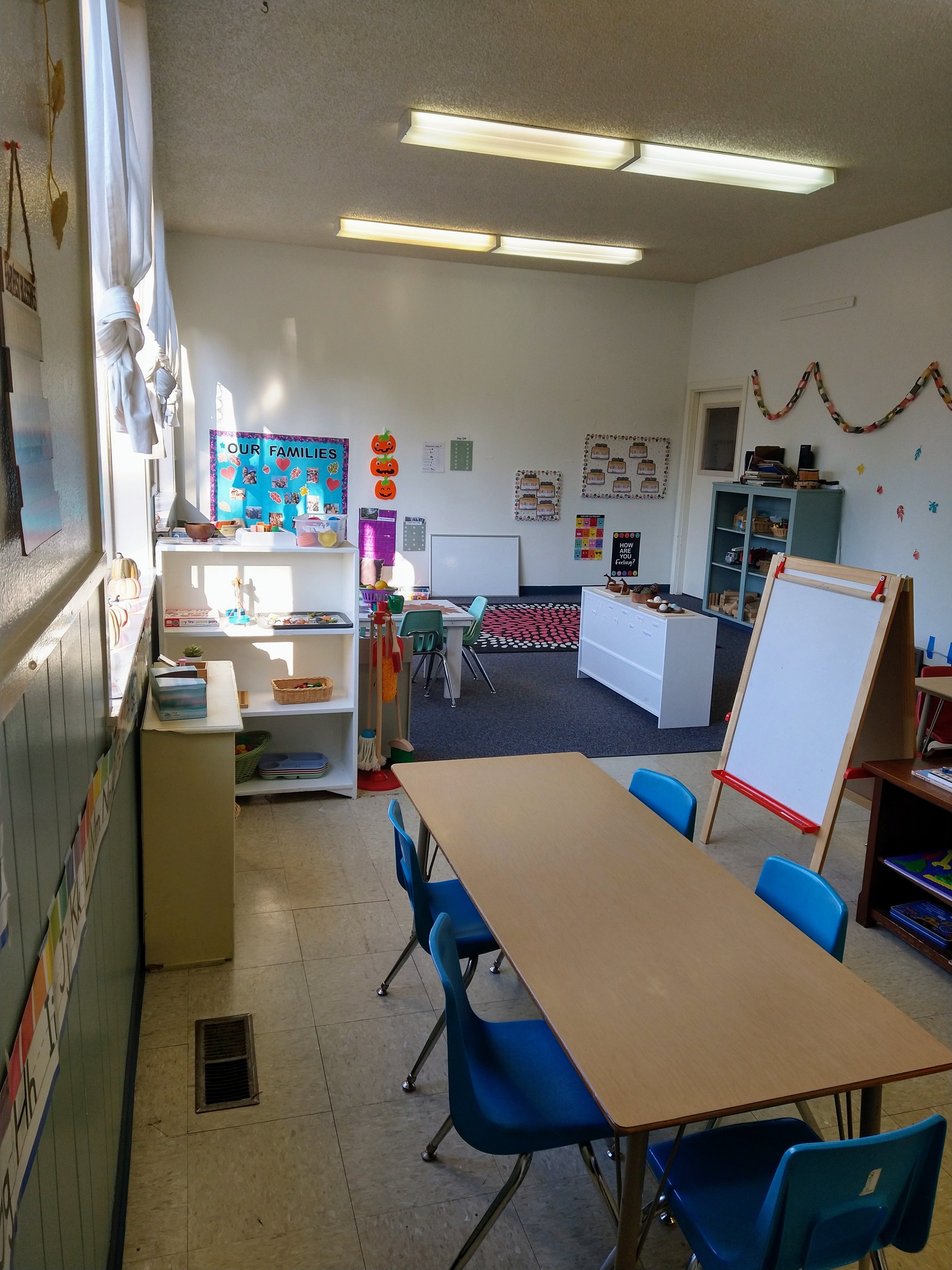  An open classroom with light coming through large windows, a toddler table, and an art easel. 