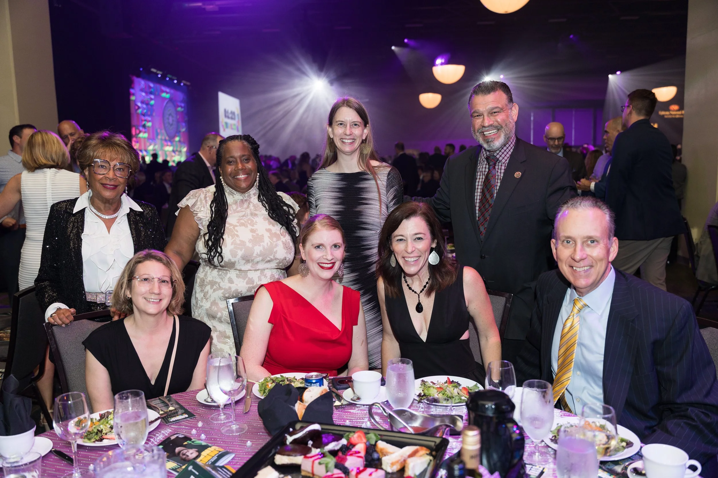 Group of eight diverse people at a banquet table, smiling, with plates of food, drinks, and desserts, in a large event hall with purple lighting, chandeliers, and other guests in the background.