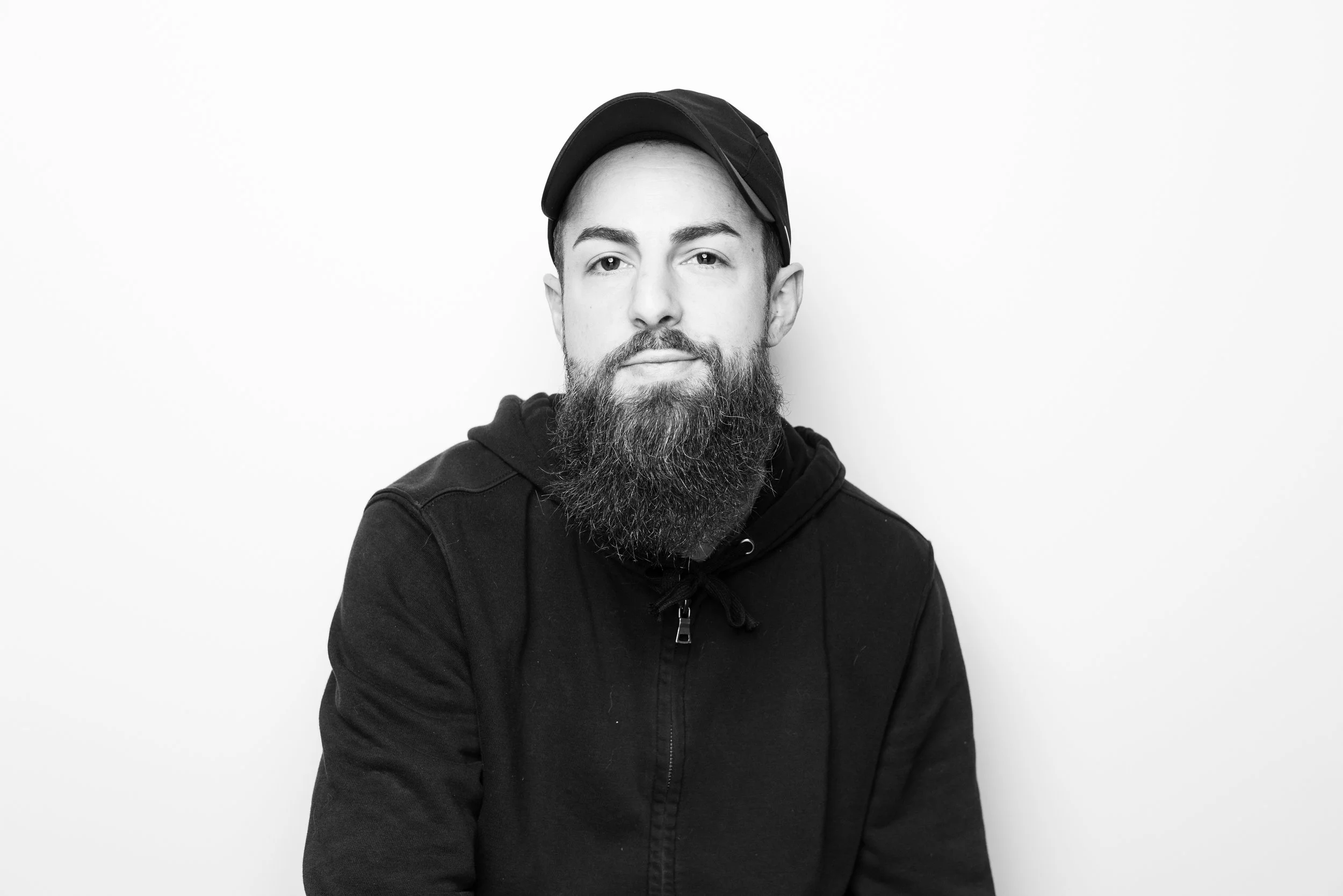 Black and white portrait of a young man with a beard, wearing a cap and hoodie, against a plain white background.