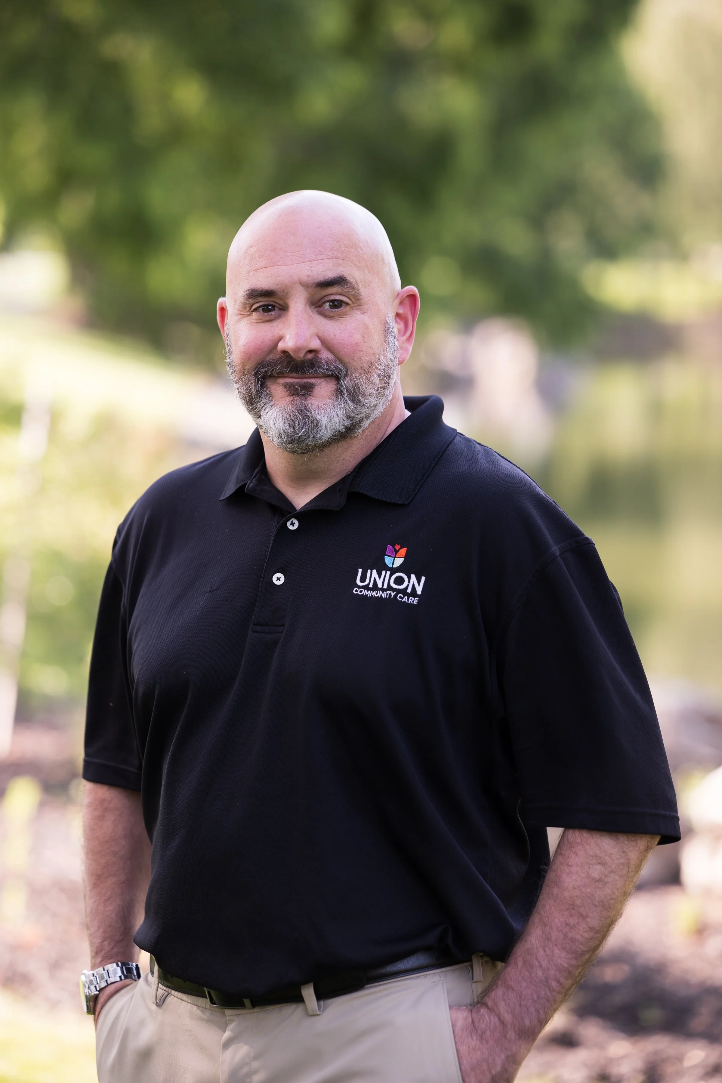 A bald man with a beard, wearing a black polo shirt with a 'Union Community Care' logo, standing outdoors with a blurred green background.