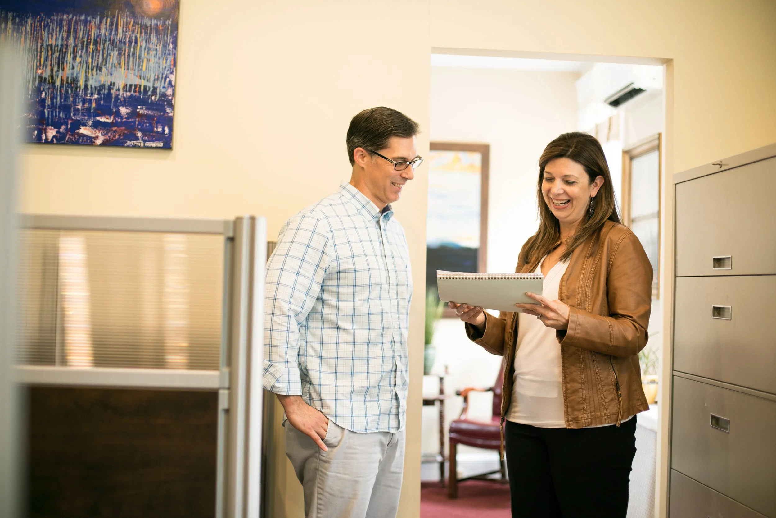 A man and woman having a conversation in an office, the woman is holding a notepad and laughing.