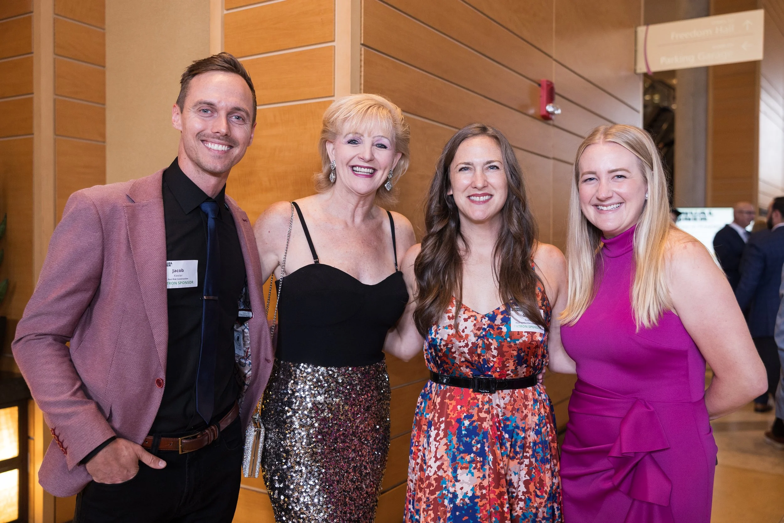 Four people at an event, smiling for the camera. They are standing indoors against a wooden wall. The man on the left is wearing a maroon blazer, black shirt, and tie. The second person is a woman with blonde hair, wearing a black top and a sequin skirt. The third person is a woman with long brown hair, wearing a multicolored patterned dress. The fourth person is a woman with long blonde hair, wearing a pink sleeveless dress. There are other people in the background, slightly out of focus.