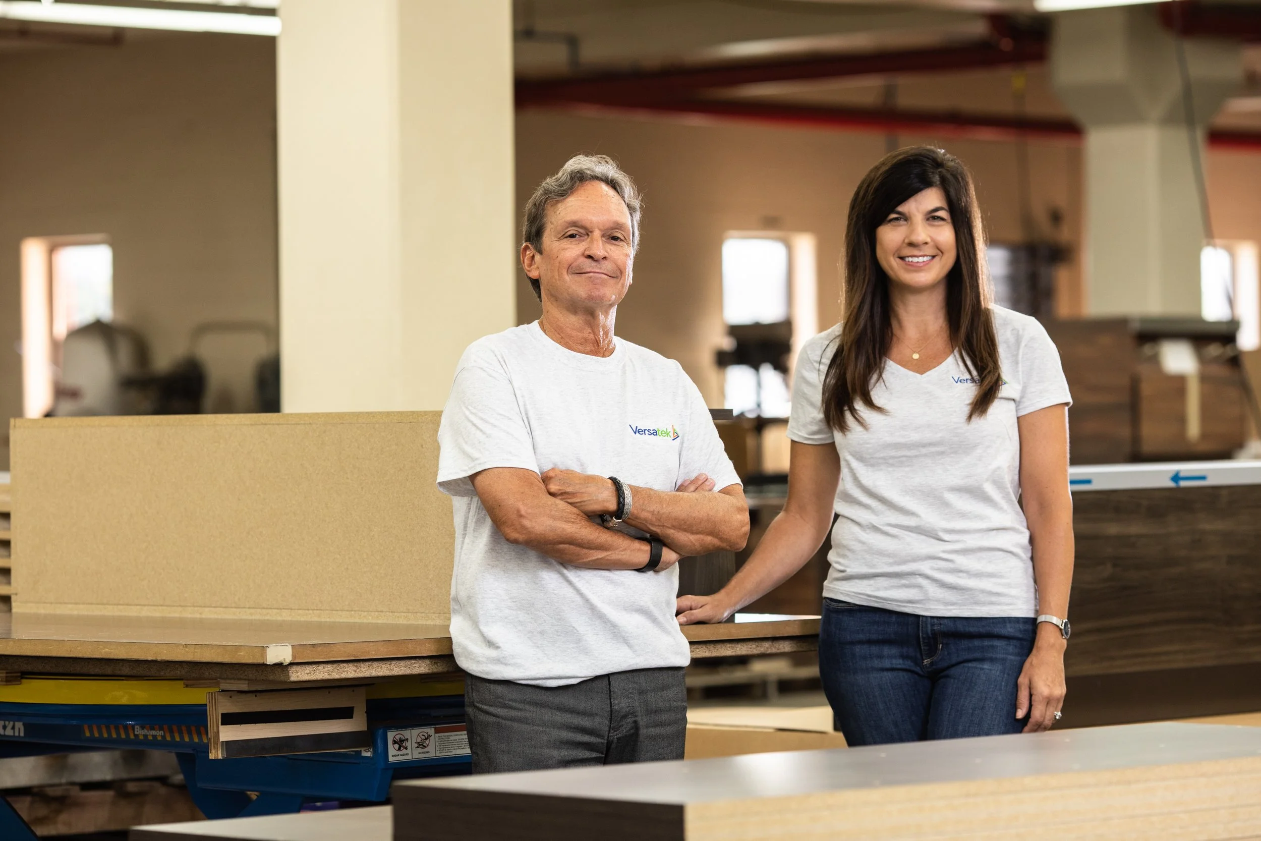 Two people, a man and a woman, standing in a woodworking shop, both wearing gray T-shirts with a logo, smiling at the camera.