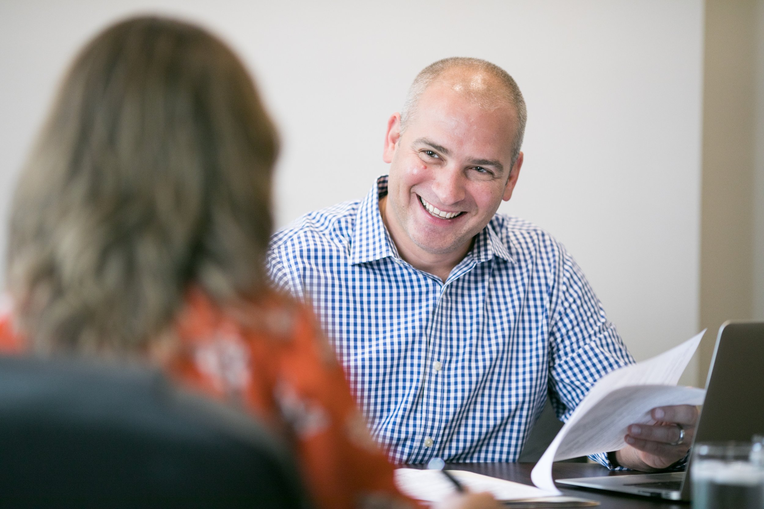 A man smiling and holding papers in a meeting with a woman whose back is to the camera.
