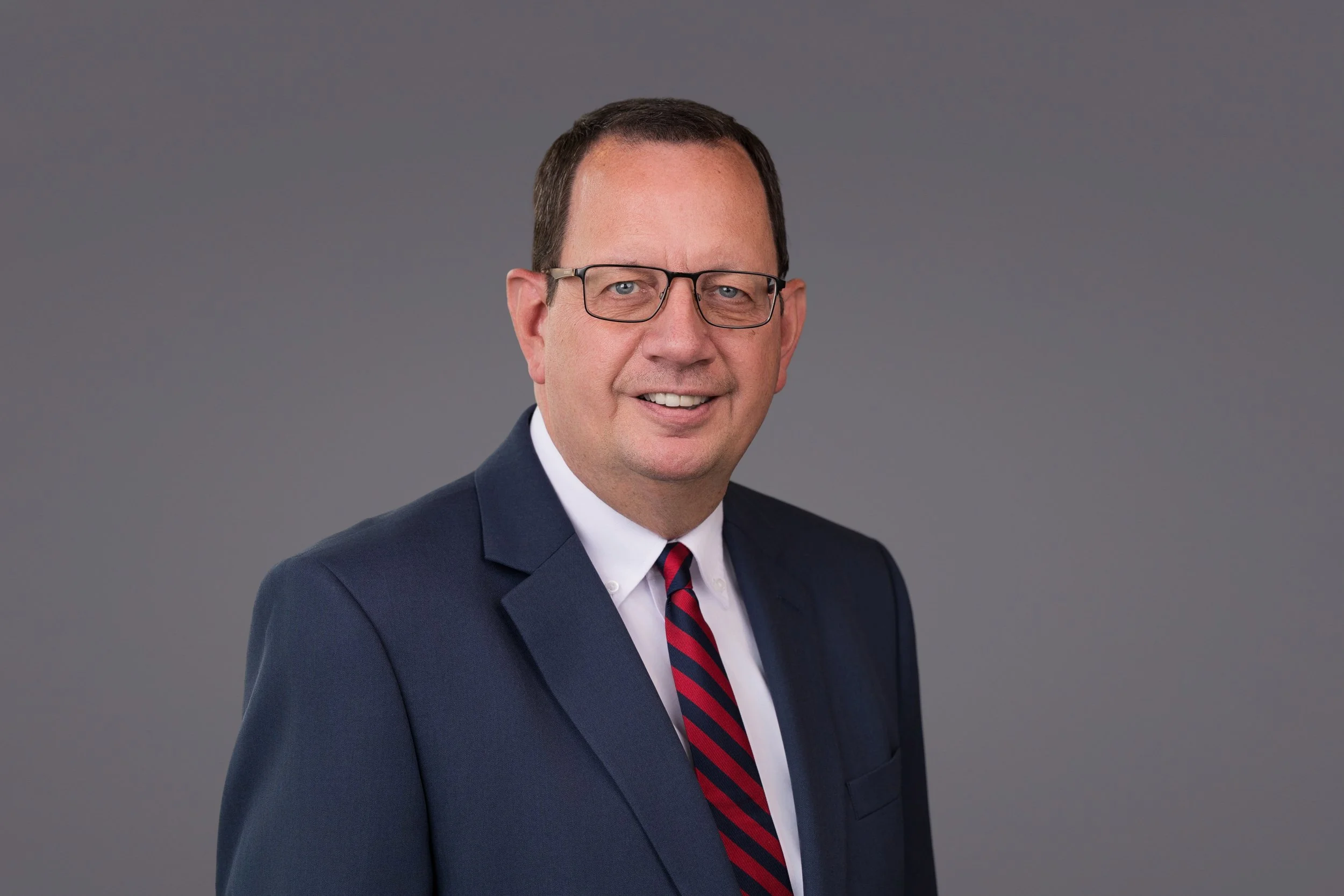 Professional headshot of a middle-aged man wearing a navy suit, white shirt, and striped red and navy tie, smiling against a plain gray background.