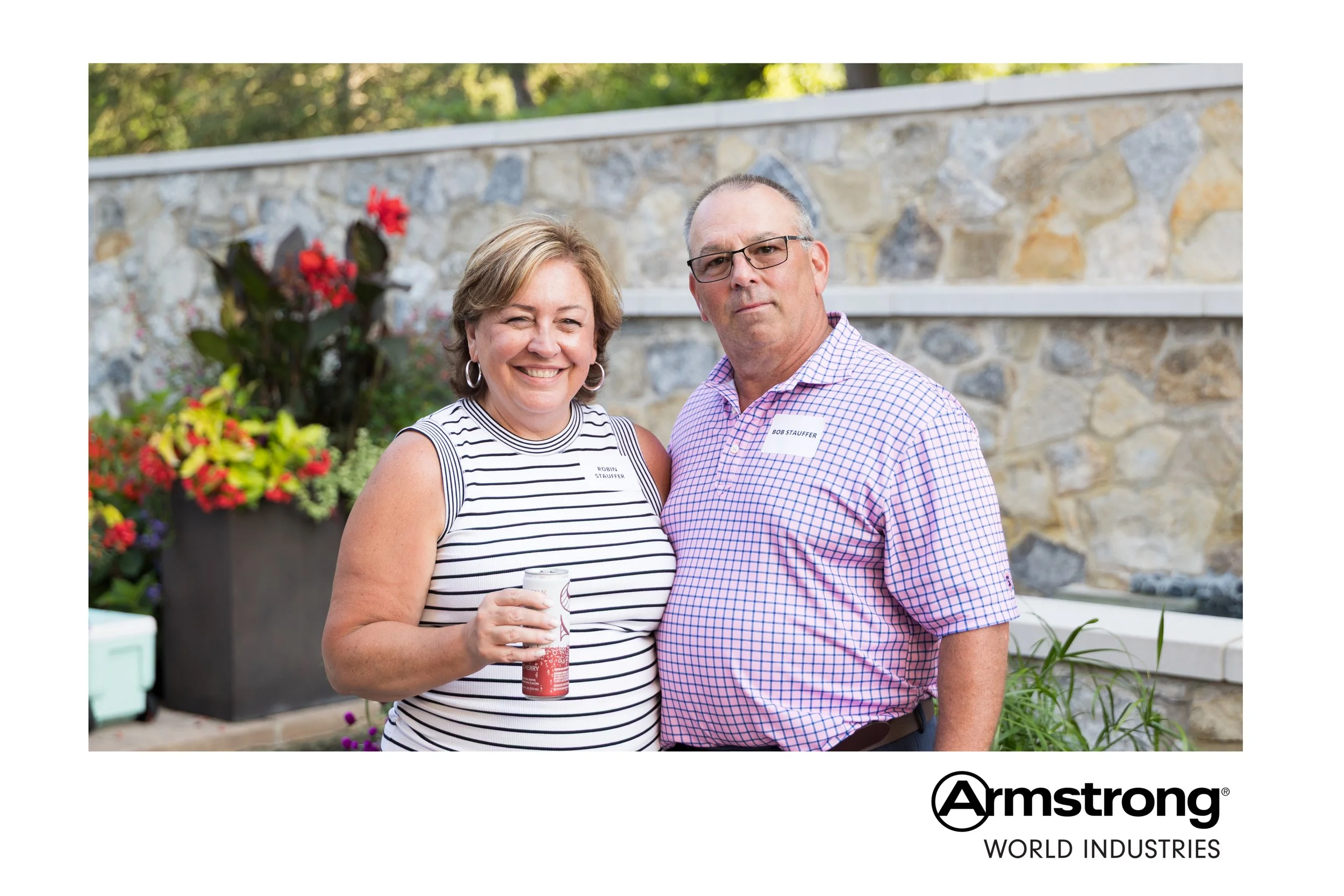 A smiling woman holding a drink standing next to a serious-looking man outdoors with a stone wall and flowers in the background.
