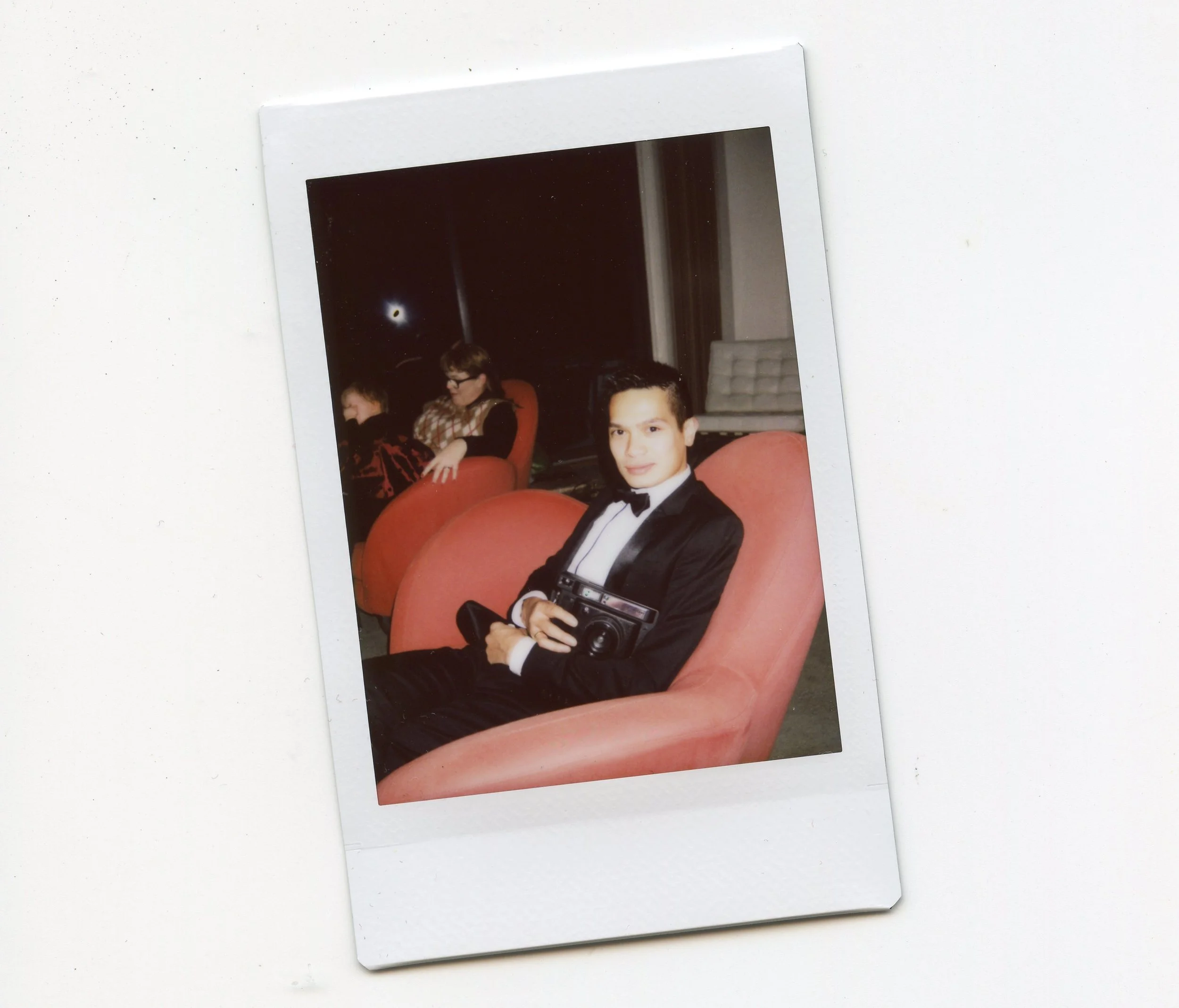 A young man in a tuxedo with a bow tie sitting on a pink chair, holding a vintage camera, with two children sitting on similar chairs in the background.