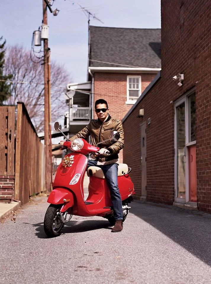 A man riding a red scooter in an alley between brick buildings, wearing sunglasses and a brown jacket.