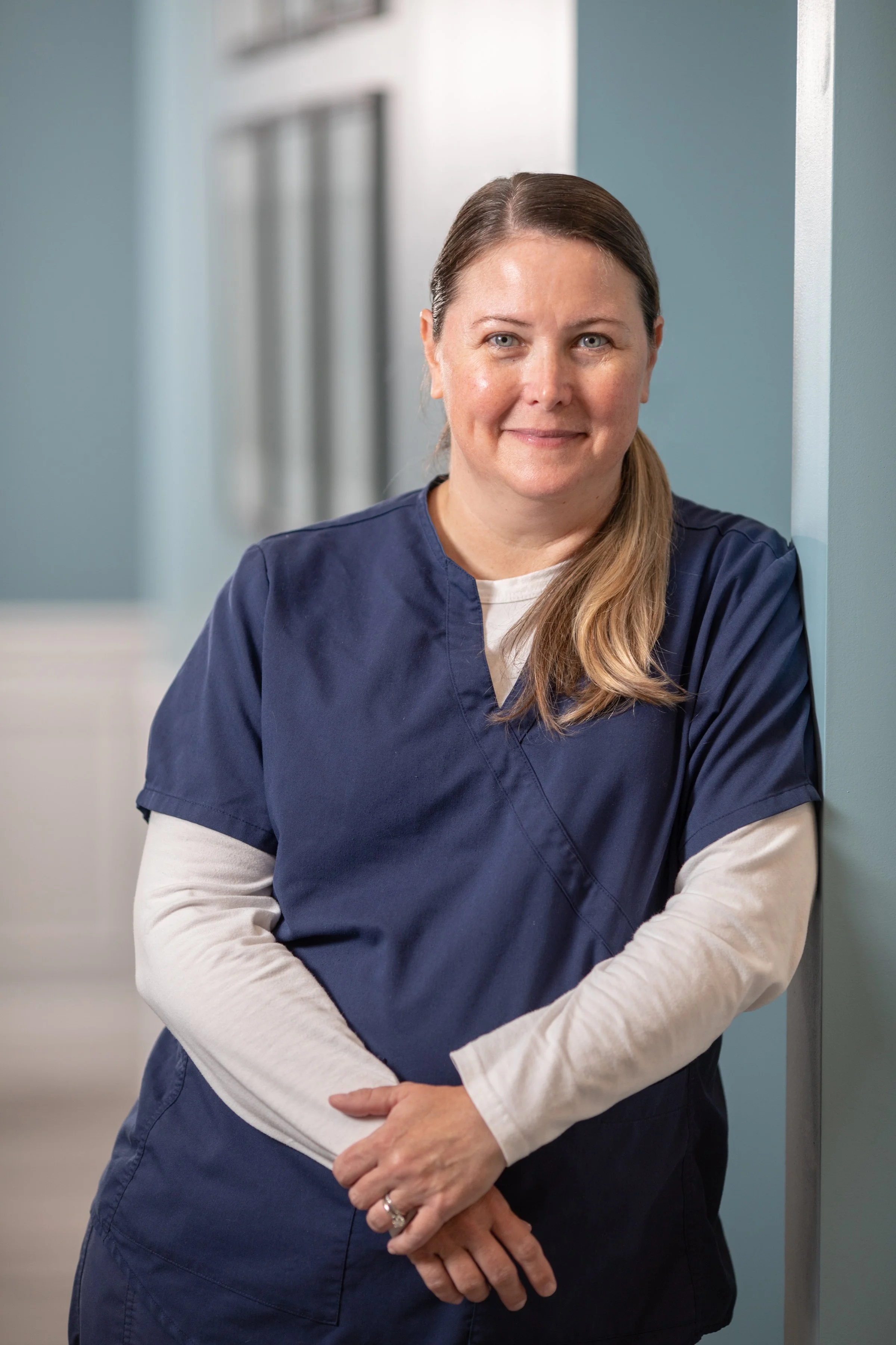 A healthcare worker wearing navy scrubs and a white long sleeve shirt standing against a teal wall in a medical facility.