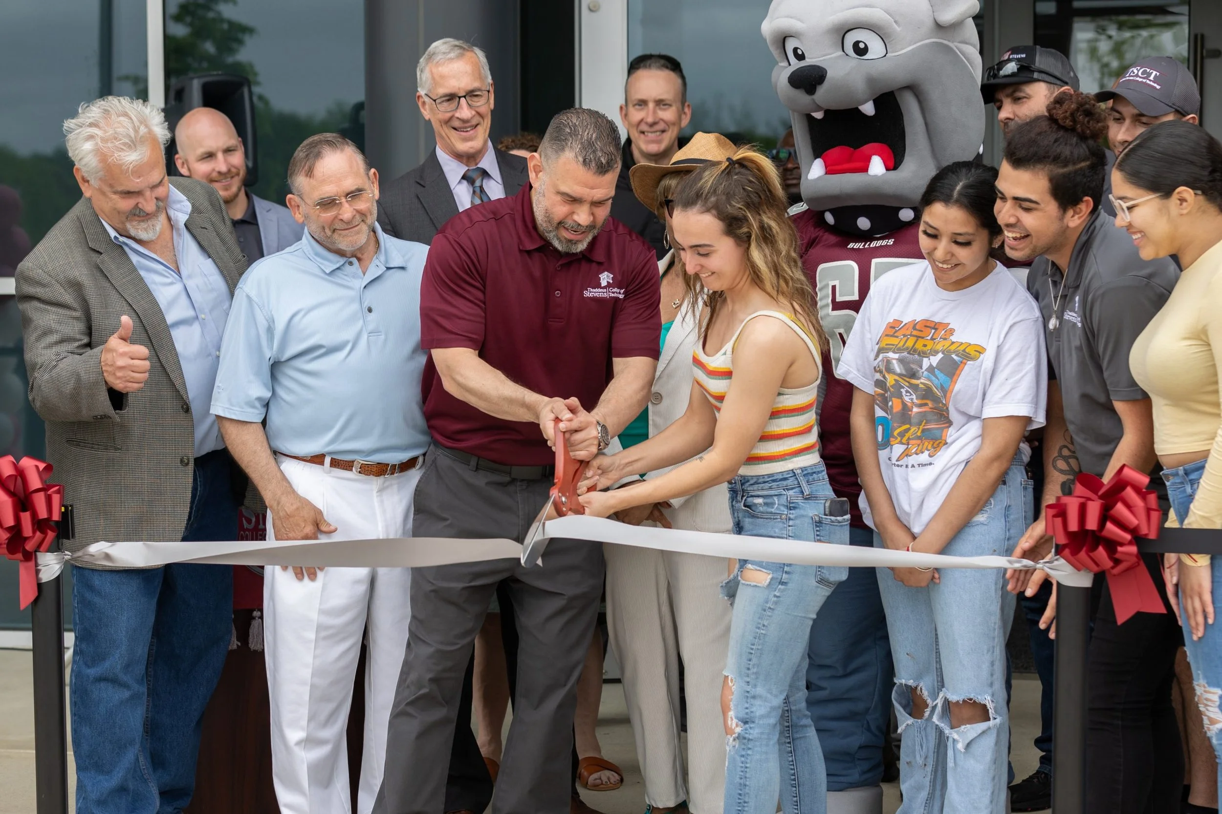 Group of people at a ribbon-cutting event outside a building, with a mascot in a bulldog costume in the background, celebrating the opening of a new facility.