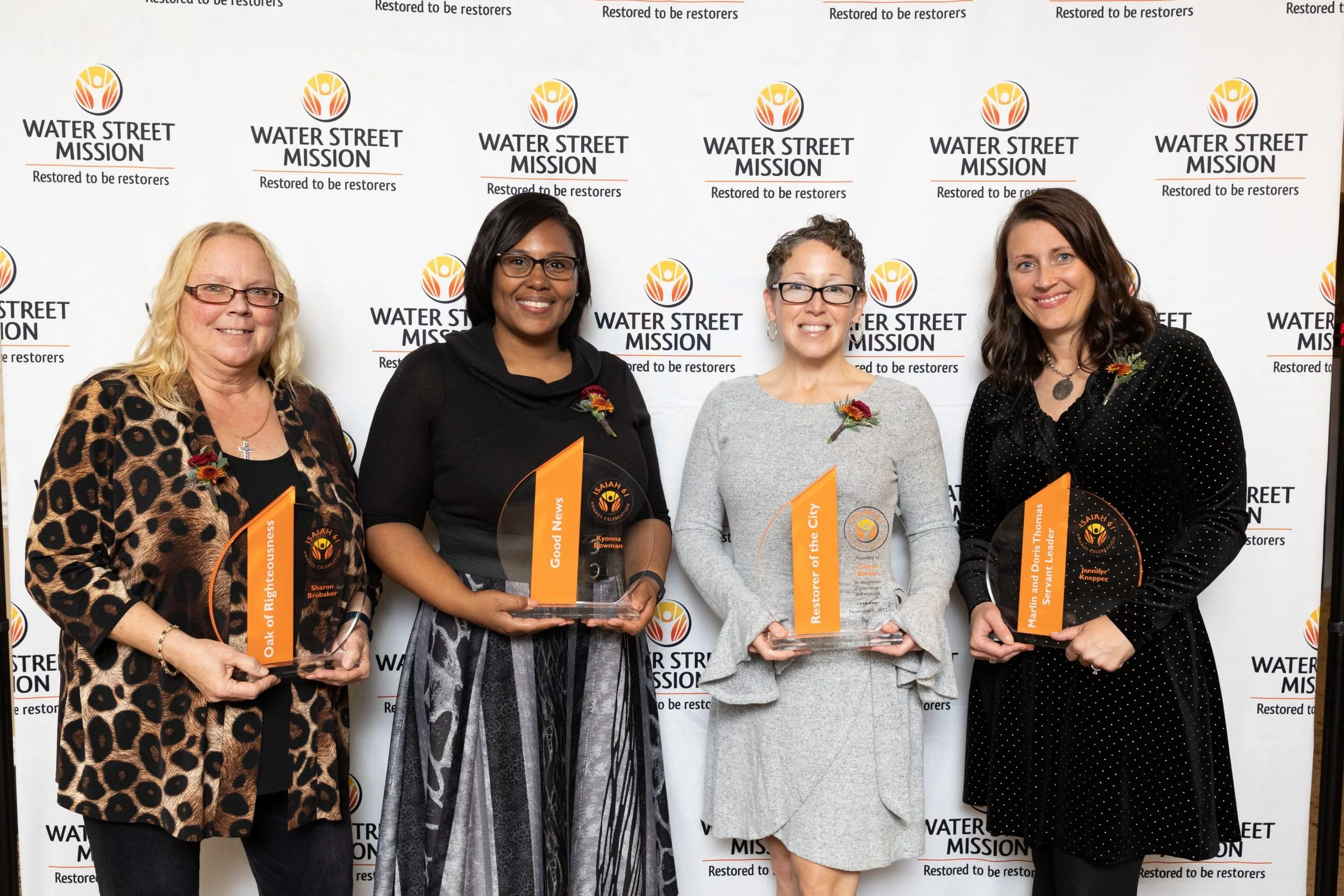 Four women standing in front of a Water Street Mission backdrop, holding awards, wearing floral pins, dressed in business casual attire.
