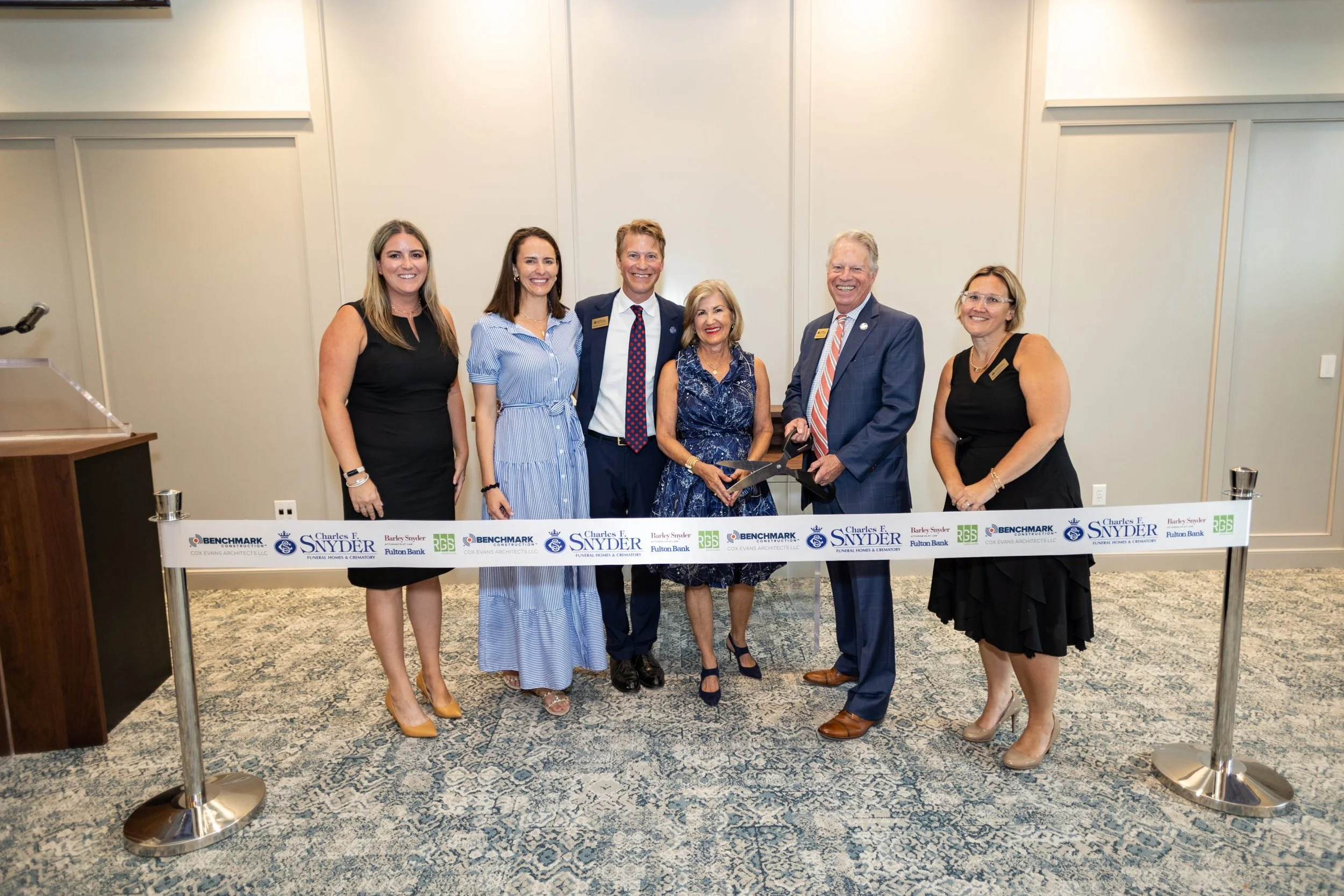 Group of six people standing behind a ribbon at a ribbon-cutting ceremony in a conference room.