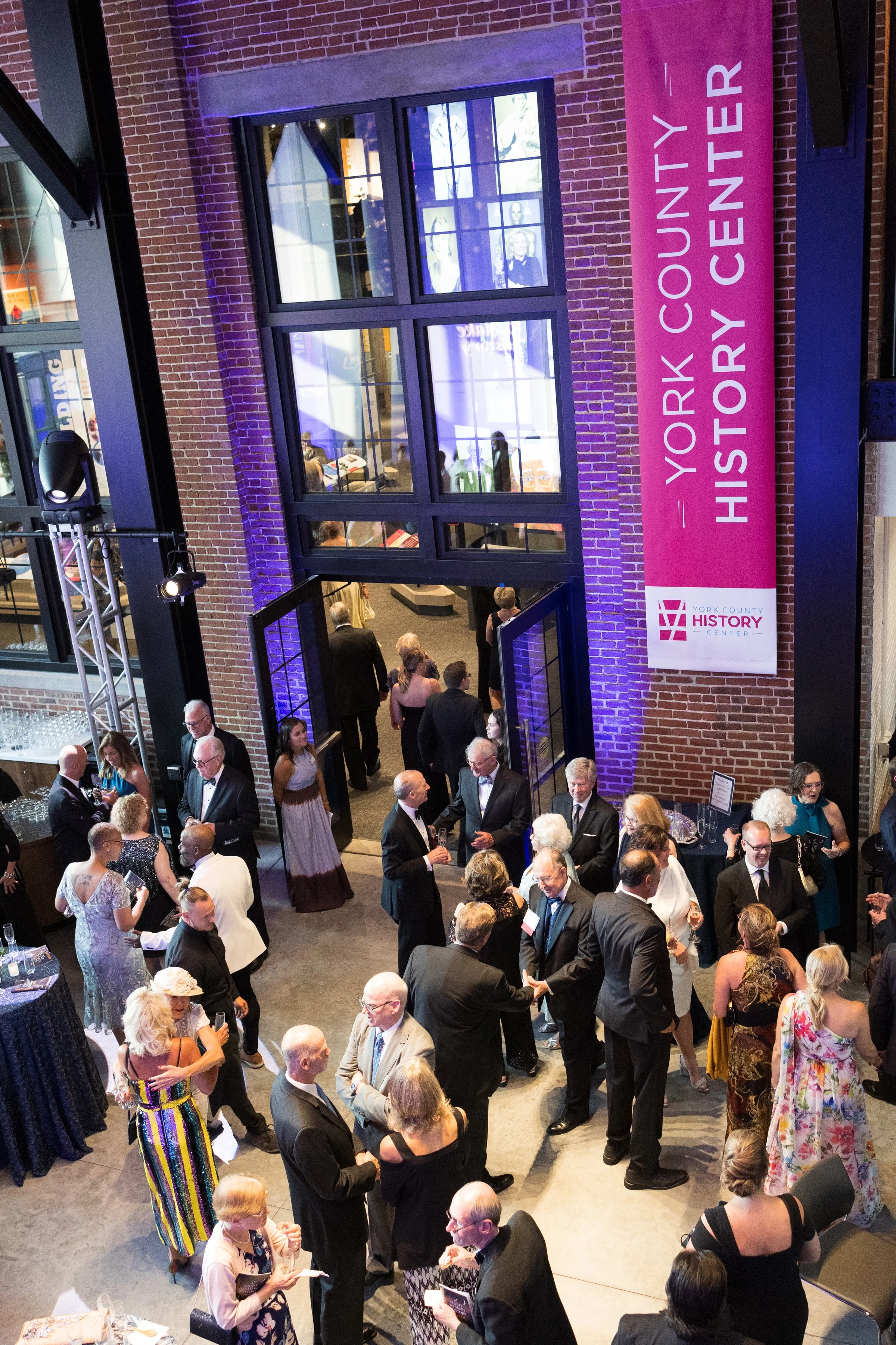A large group of people dressed in formal attire attending a social event at the York County History Center. The event is happening inside a renovated brick building with a sign reading "York County History Center." Some guests are mingling and chatting, while others are walking through a double glass door. There are tables with drinks and glasses, and the event appears lively and well-attended.