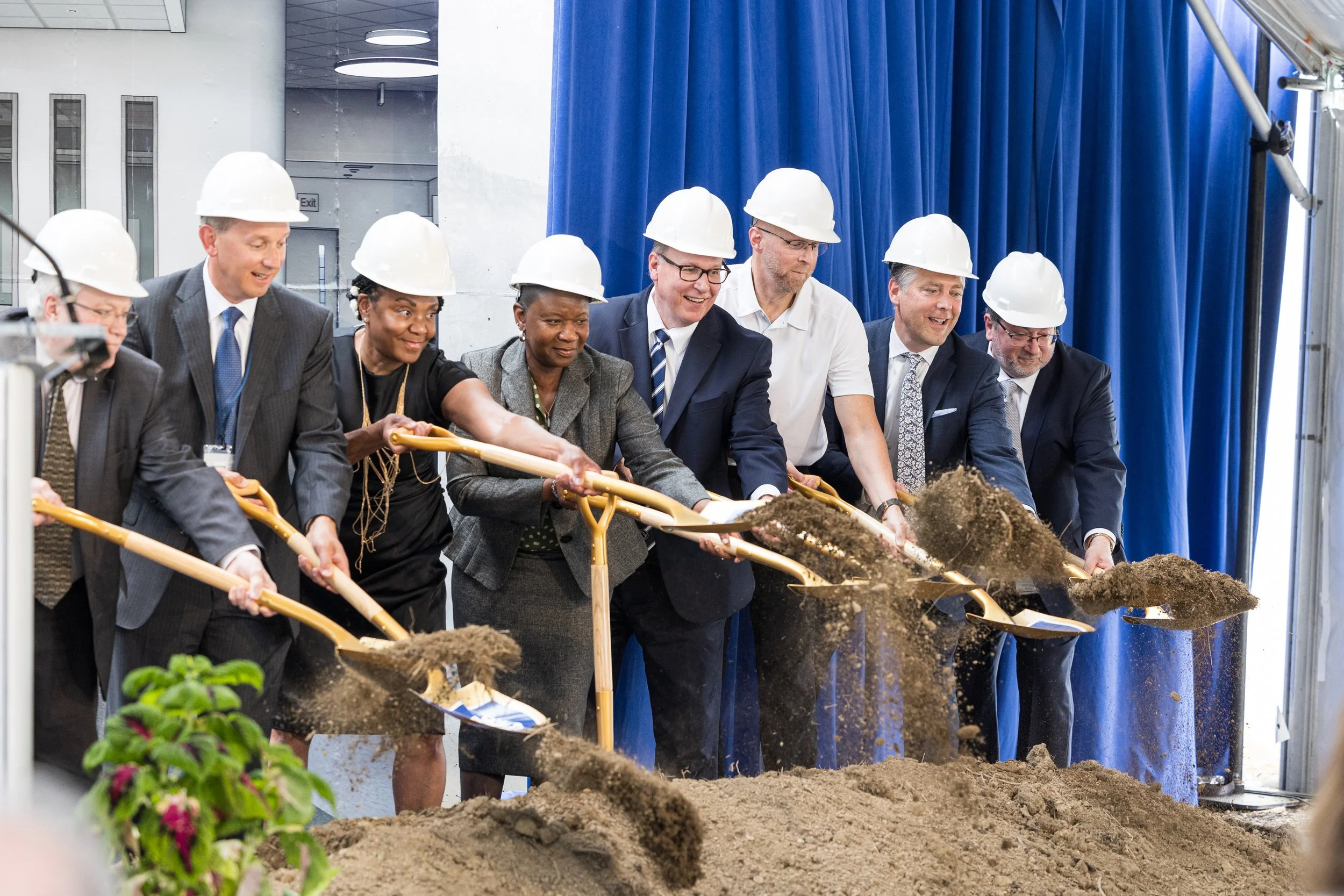 People in suits and hard hats at a groundbreaking ceremony, shoveling dirt into a pile.
