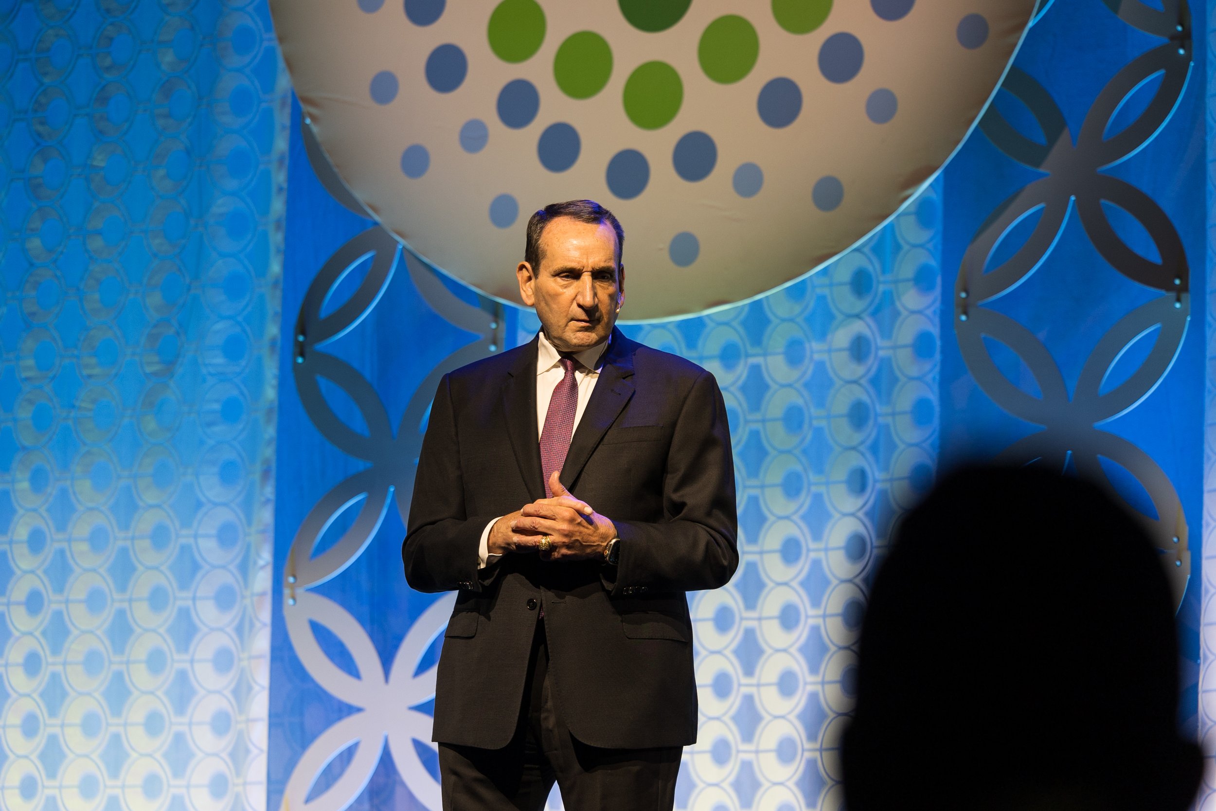 A man in a black suit, white shirt, and purple tie standing on a stage with a blue-themed geometric backdrop, looking slightly to the side with a serious expression.