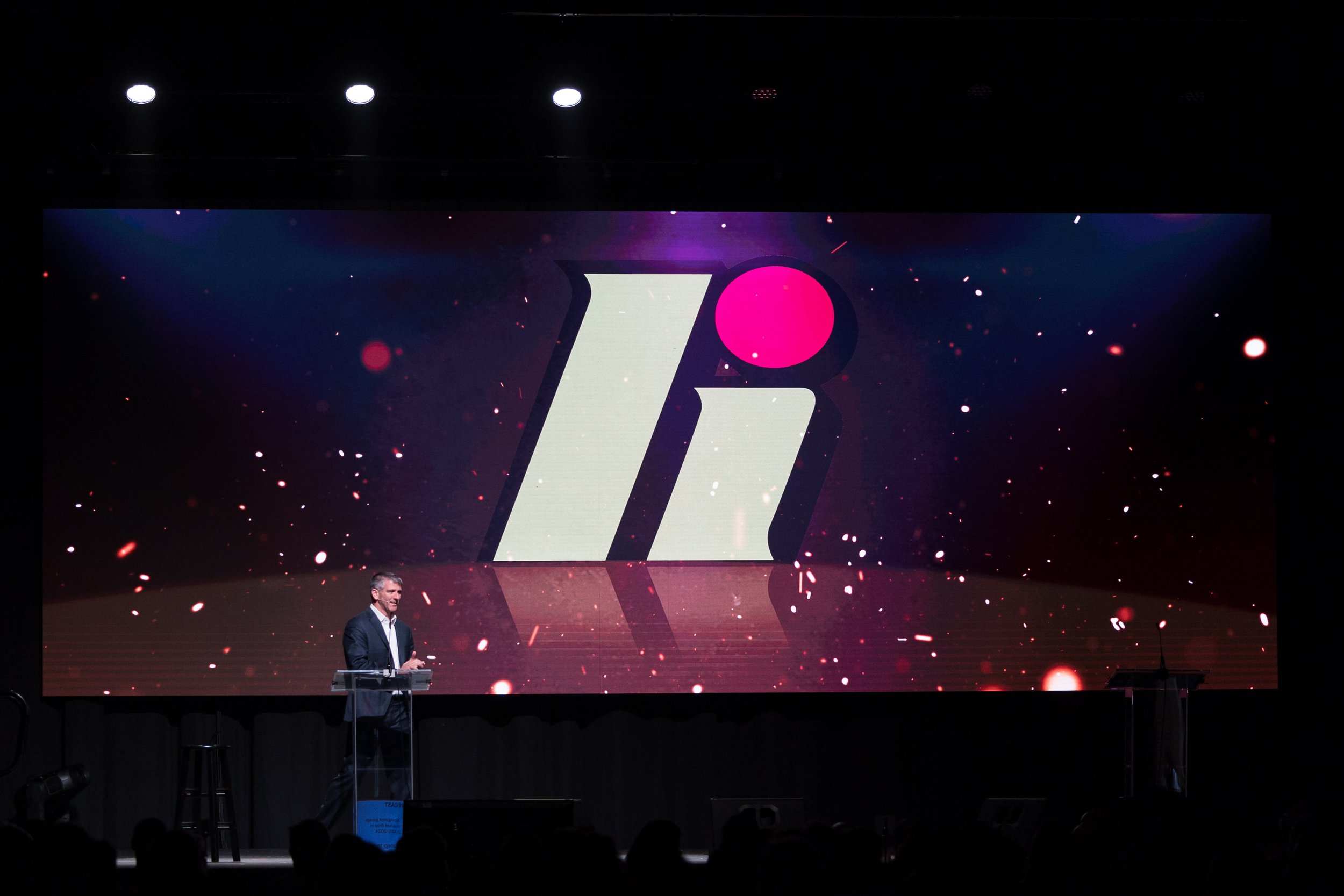 A man standing at a clear podium on a dark stage with a large screen behind him displaying a logo with a white lowercase 'i' and a pink dot above it, set against a black background with red and white particle effects.